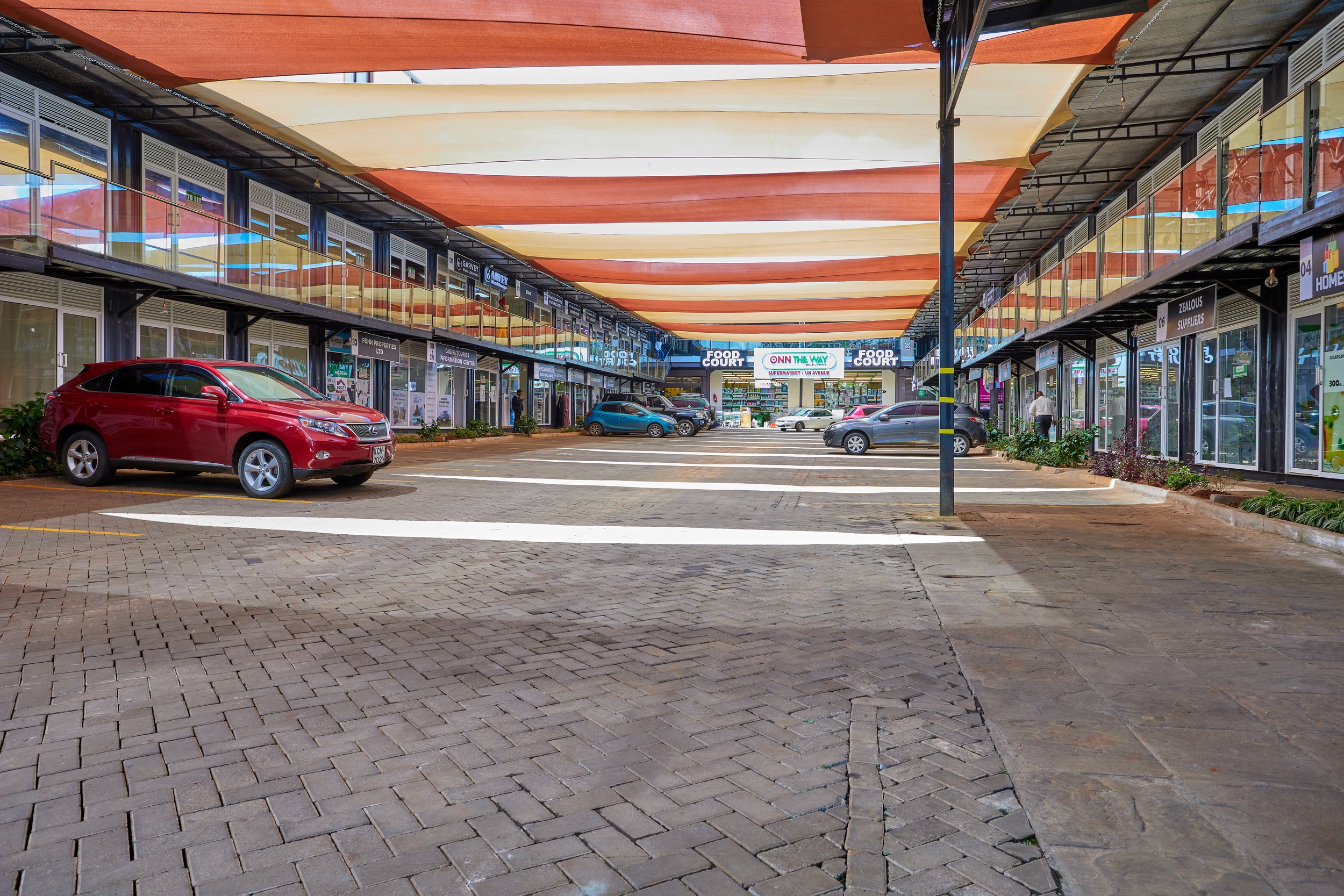 Outdoor shopping center parking lot with cars parked, storefronts on sides, and a sign that reads 'Food Court' and 'On The Way' in the background.
