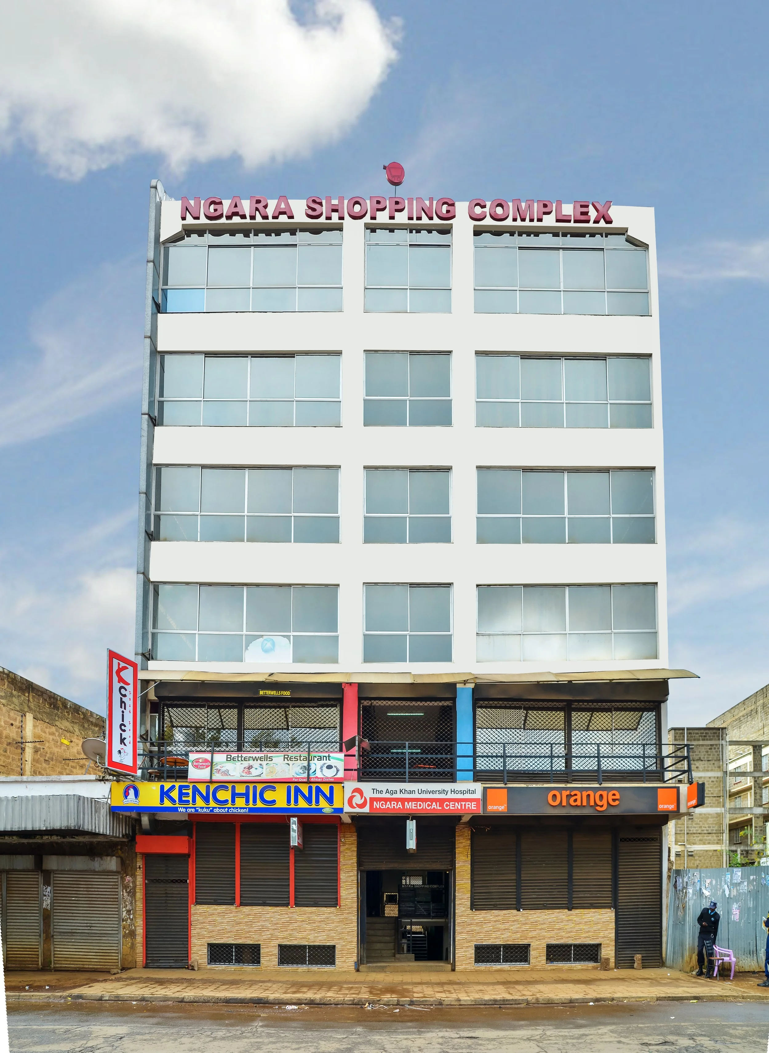 Multi-story building with signs for Ngara Shopping Complex, Kenchic Inn, Ngara Medical Centre, orange store, and a restaurant on the second floor. Bright and modern architecture with a blue sky background.