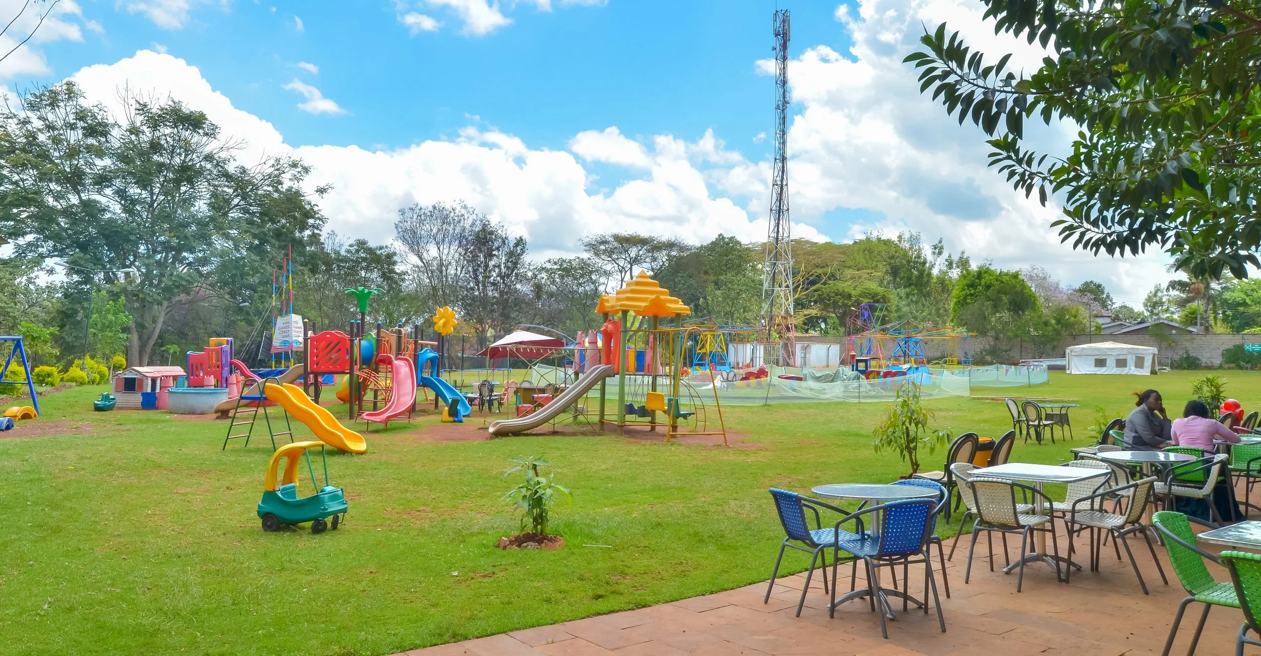 Children's playground with slides, swings, and colorful play structures in a park, with some adults seated at outdoor tables nearby.