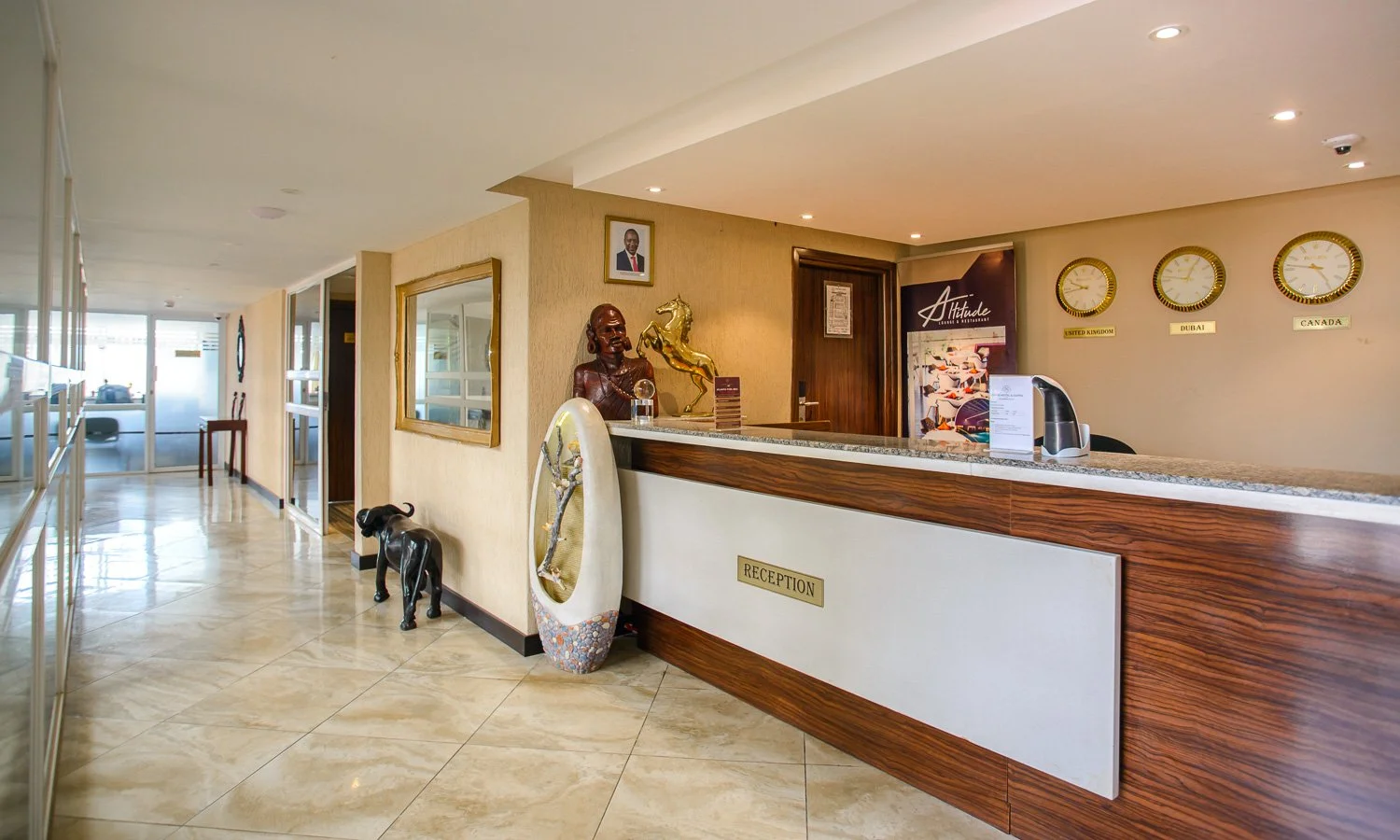 Hotel reception desk with a marble countertop, decorative sculptures, a framed photo, and 3 wall clocks displaying times for the United Kingdom, Dubai, and Canada. The area is well-lit with tiled floors and a glass front entrance in the background.