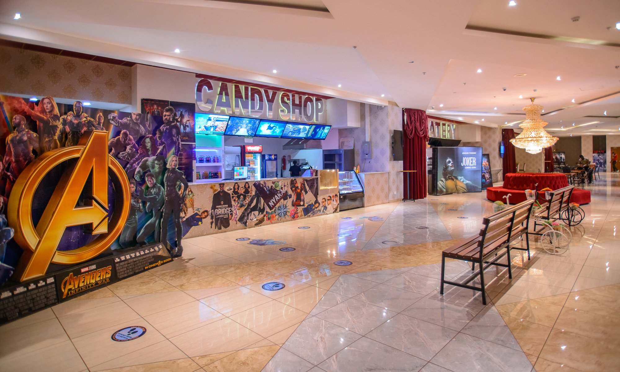 Interior of a shopping mall with a candy shop, movie posters, seating area, and a chandelier.