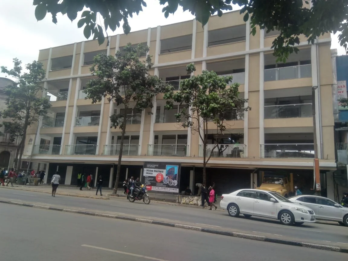 Multi-story beige building with glass balconies, pedestrians walking on the sidewalk, cars parked and a motorcycle on the street, trees in front, billboard on the building.
