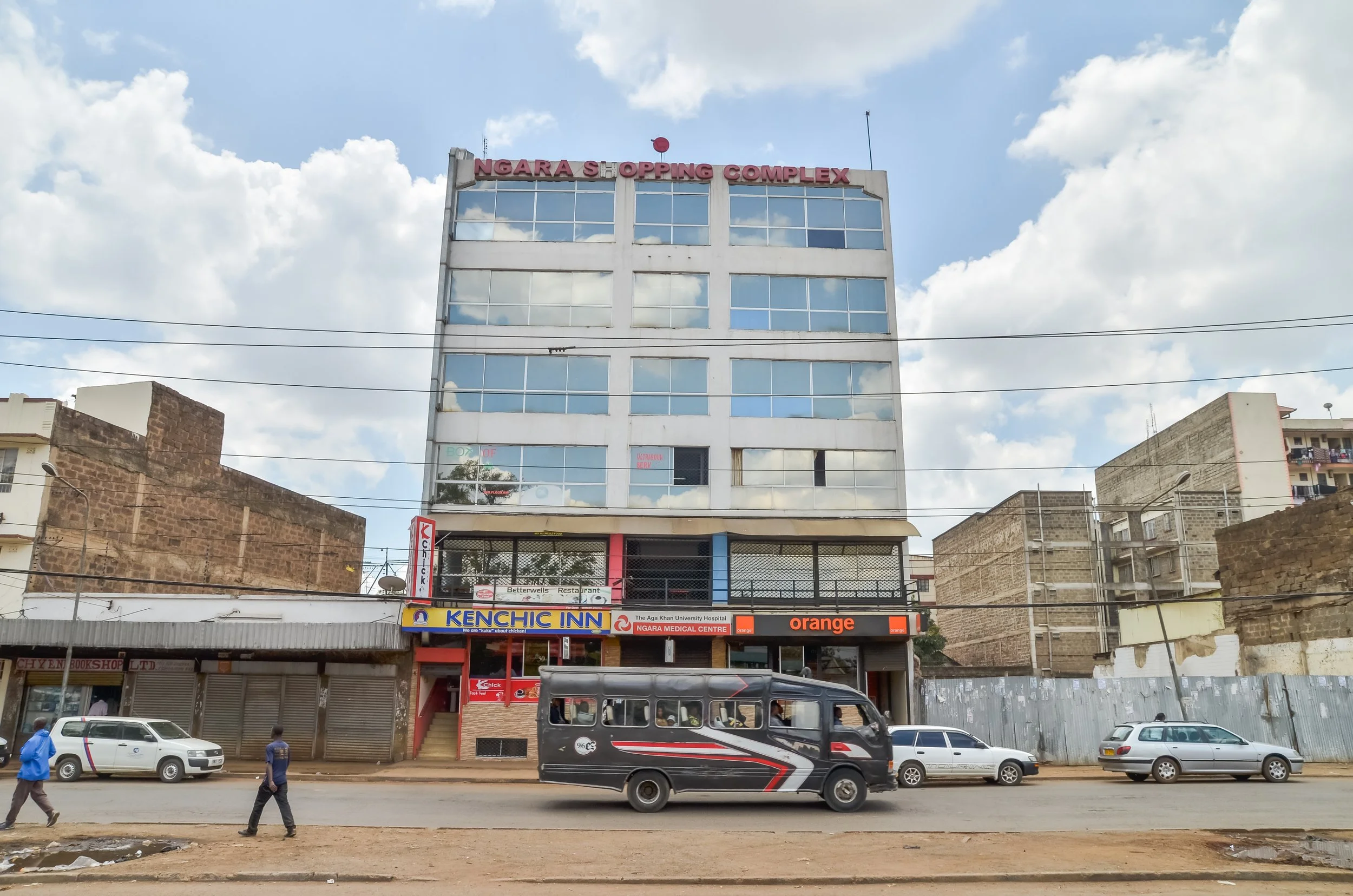 Multi-story building with the sign 'Ngara Shopping Complex' on top, storefronts including Kenchic Inn, Orange, and Ngara Medical Centre on the ground floor, a black and red matatu vehicle on the street, and pedestrians walking in front under cloudy sky.