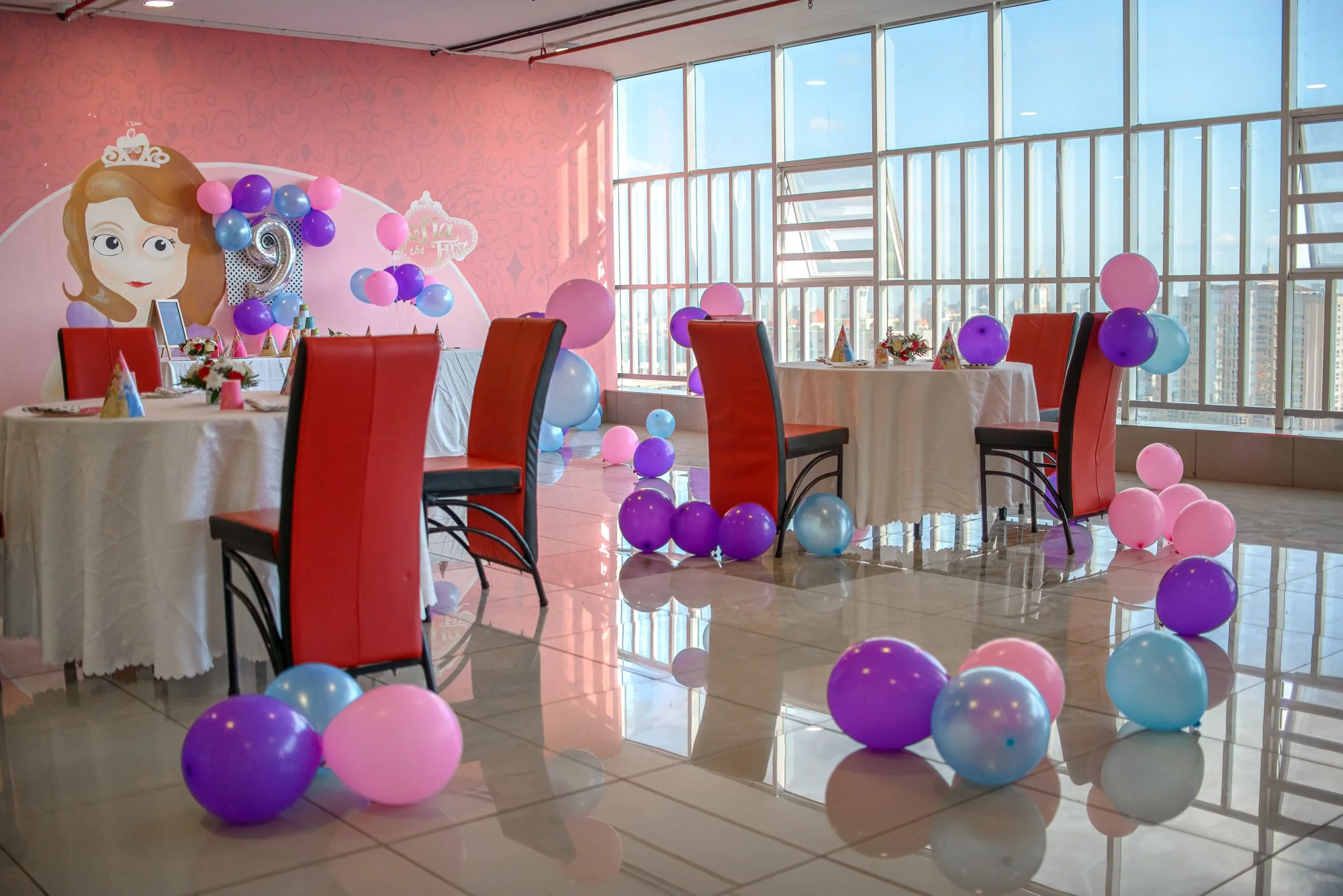 Birthday party decorated with pink, purple, and light blue balloons, featuring a princess-themed backdrop with a cartoon princess, set up with tables and red chairs in a bright room with large windows showing a city skyline.