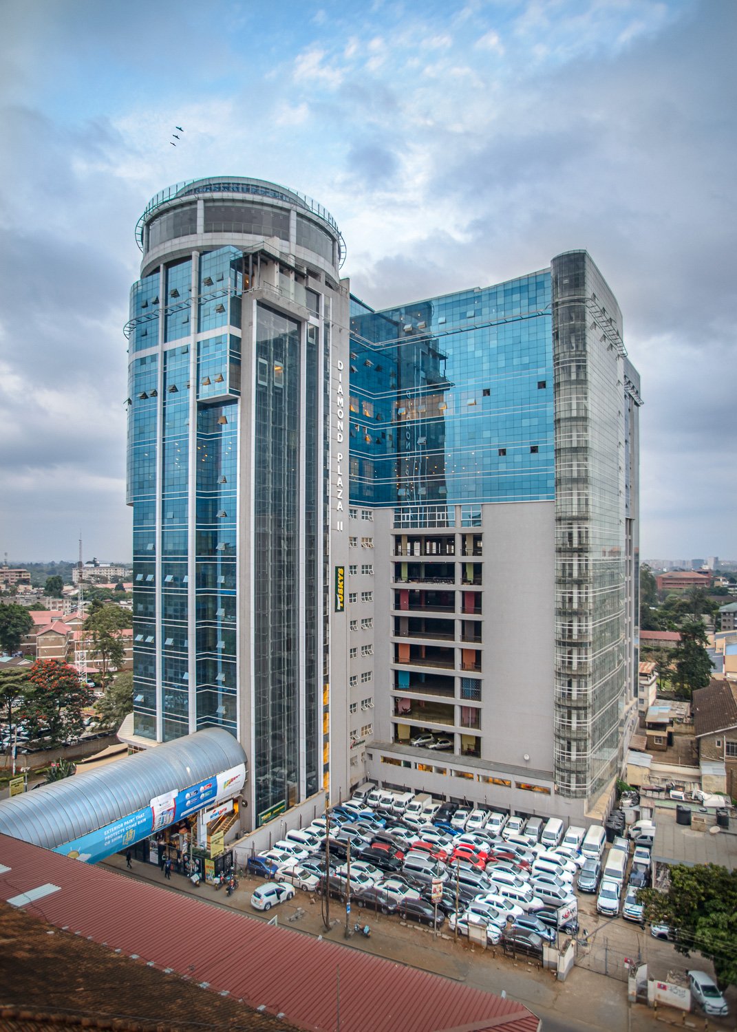 Tall modern glass office building with a parking lot filled with white cars at its base, under a partly cloudy sky.