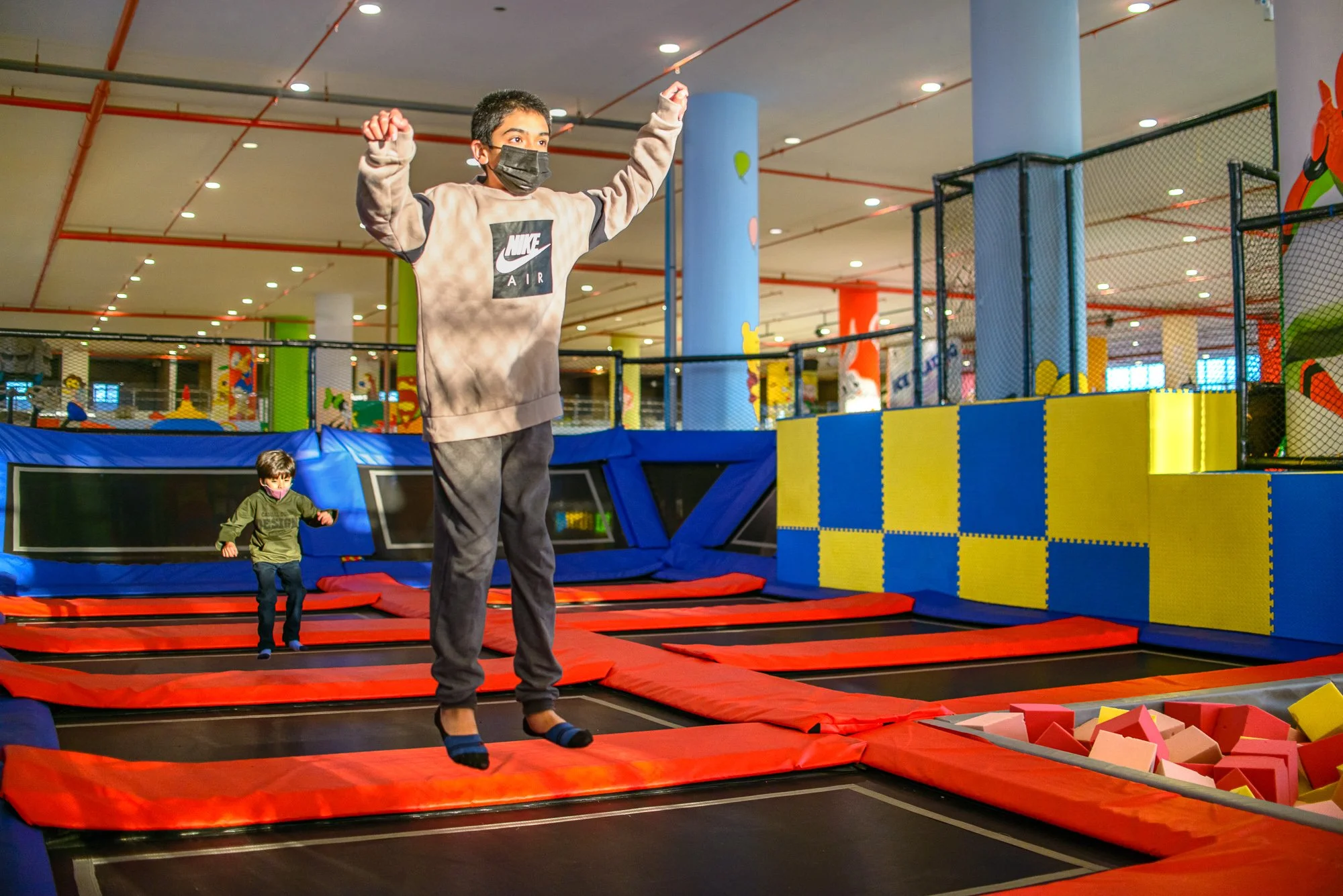 Indoor trampoline park with two children jumping and bouncing, one in the foreground wearing a hoodie and face mask, and a smaller child in the background wearing a green jacket. Brightly colored padded mats and obstacle structures are visible.