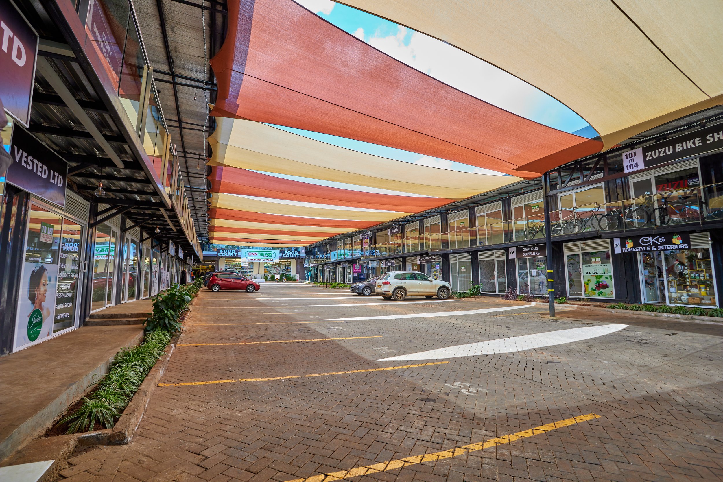Empty shopping plaza parking lot with cars parked, storefronts on both sides, and colorful fabric canopy overhead.