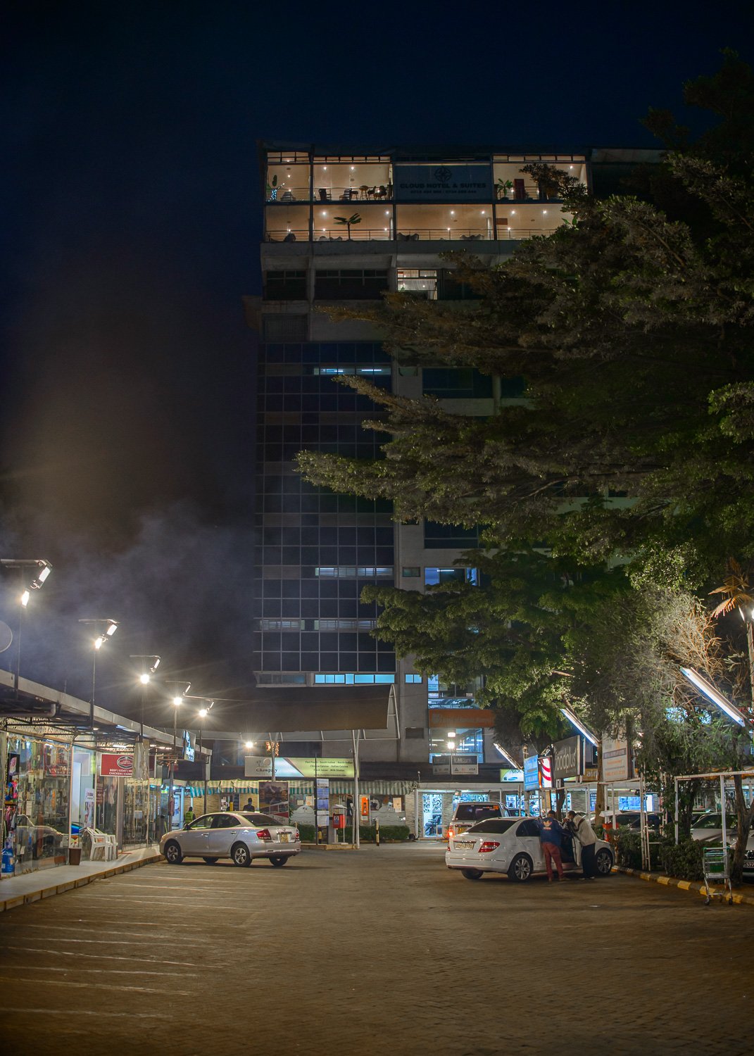 Nighttime view of a parking lot in front of a multi-story building with illuminated interior, shops on the ground floor, and a sign that reads 'Cloud Hotel & Suites'. Cars are parked, and a person is standing near an open car trunk. A large tree partially obscures the building, and streetlights illuminate the scene.