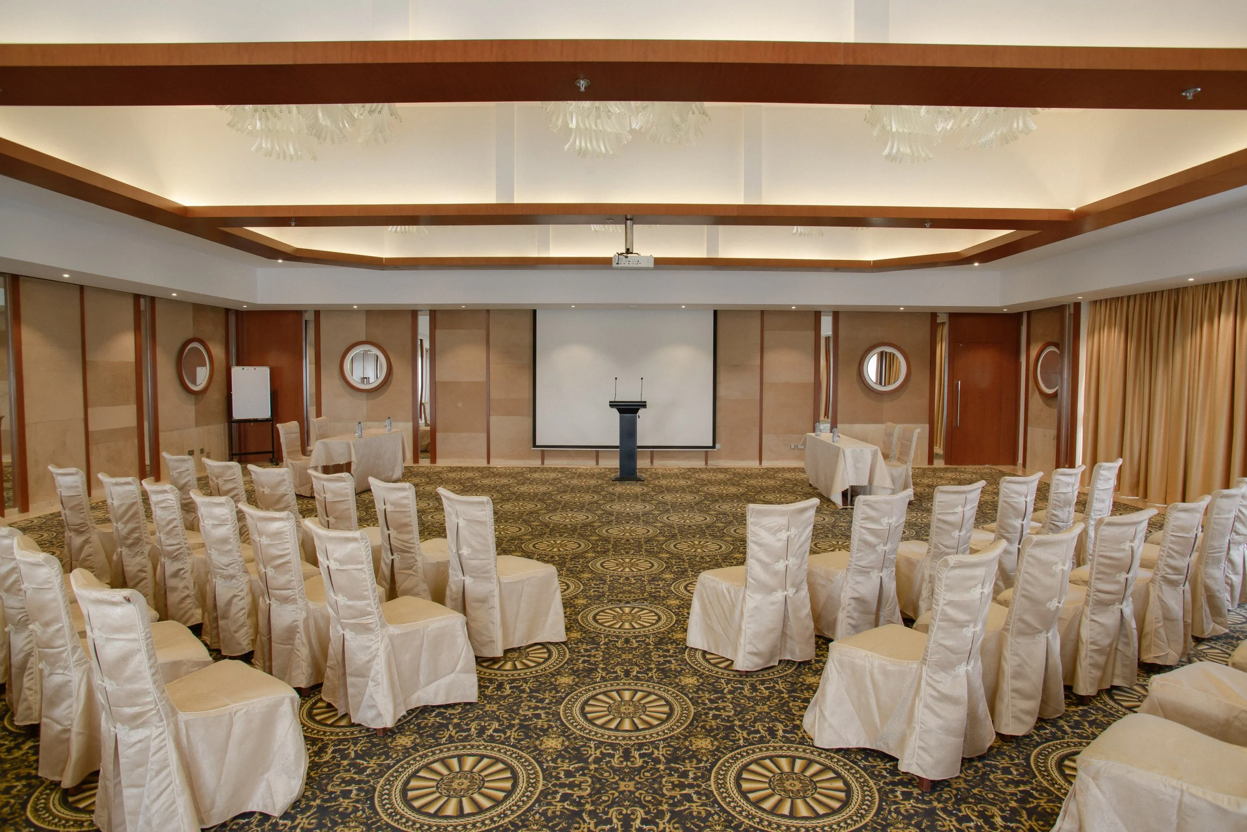 Conference room with rows of chairs covered in white cloth, facing a large screen and a podium, with a patterned carpet and beige walls.