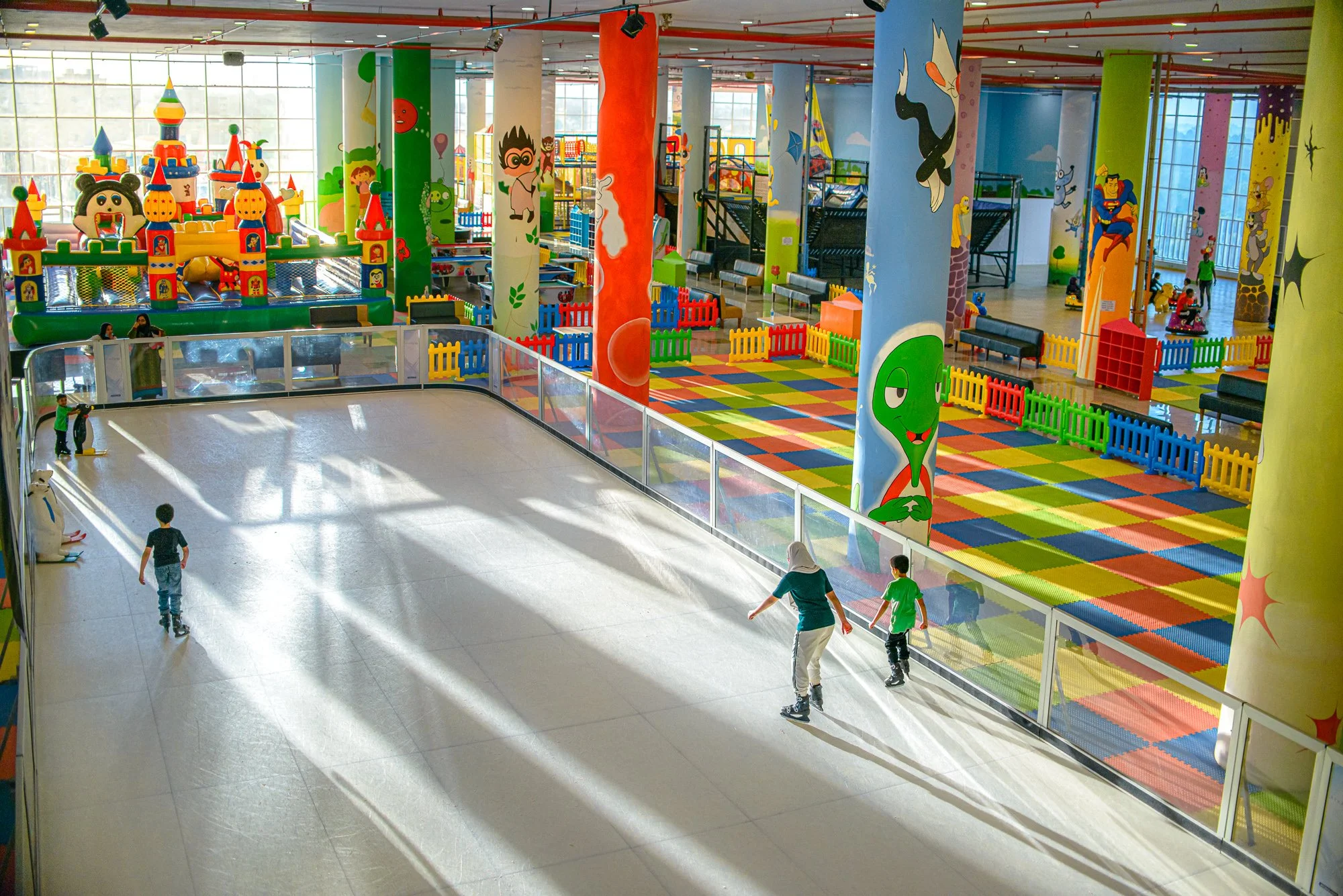 Indoor children's playground with colorful climbing structures, padded flooring, and a skating rink area with a woman and children skating.