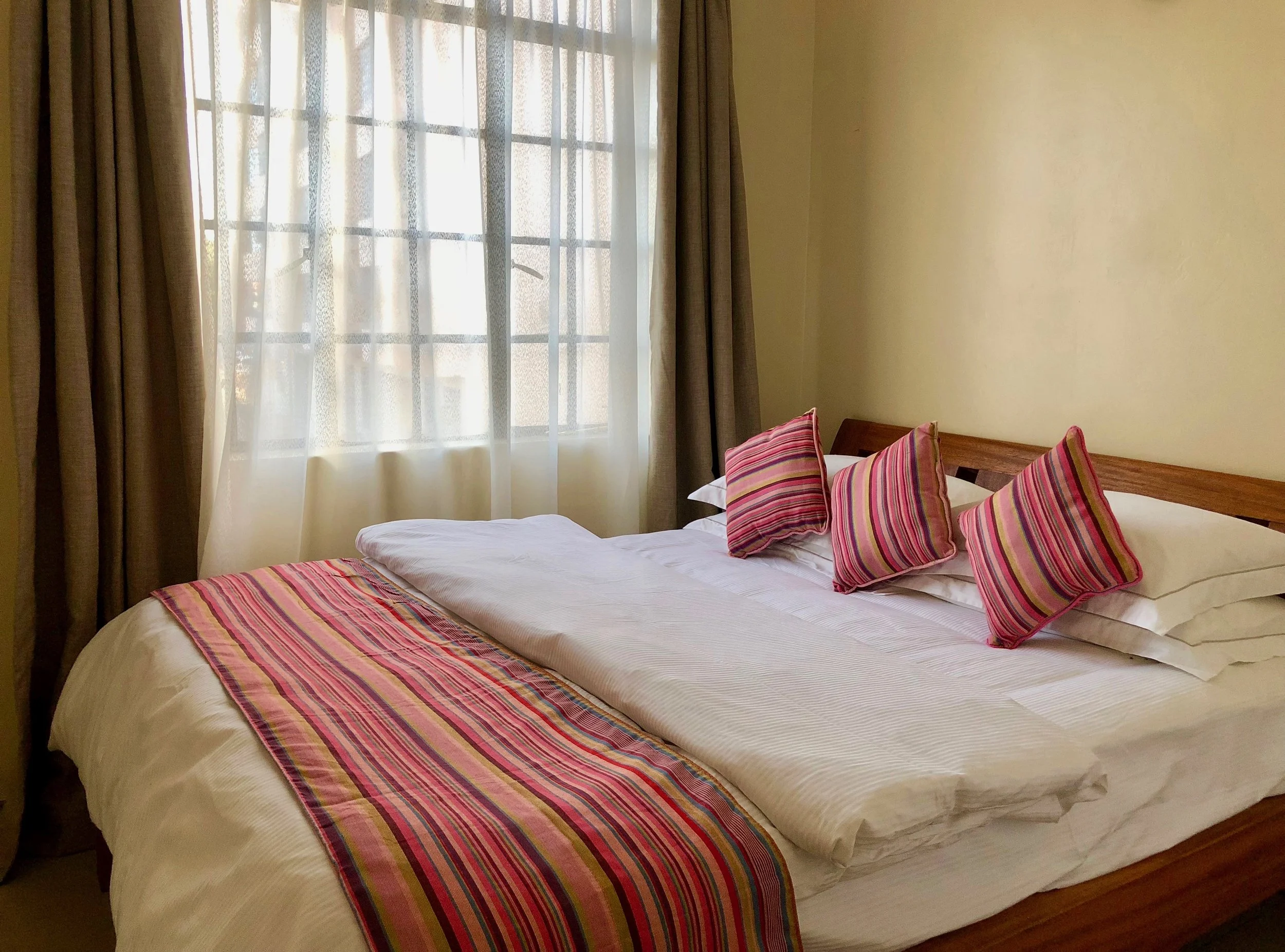 A neatly made bed with white linens, striped pink and red pillows, and a matching striped bed runner in a room with a window covered by sheer and brown curtains.
