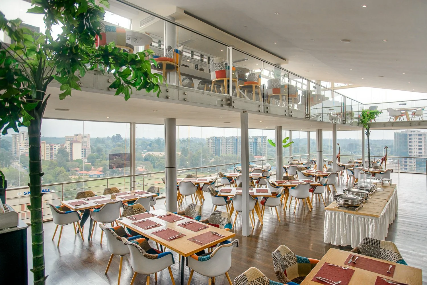 Modern restaurant with large windows, multiple tables set for dining, some with placemats and silverware, and decorative giraffe statues near the railing. Two levels with the upper balcony visible, dotted with chairs and tables, overlooking the cityscape outside.