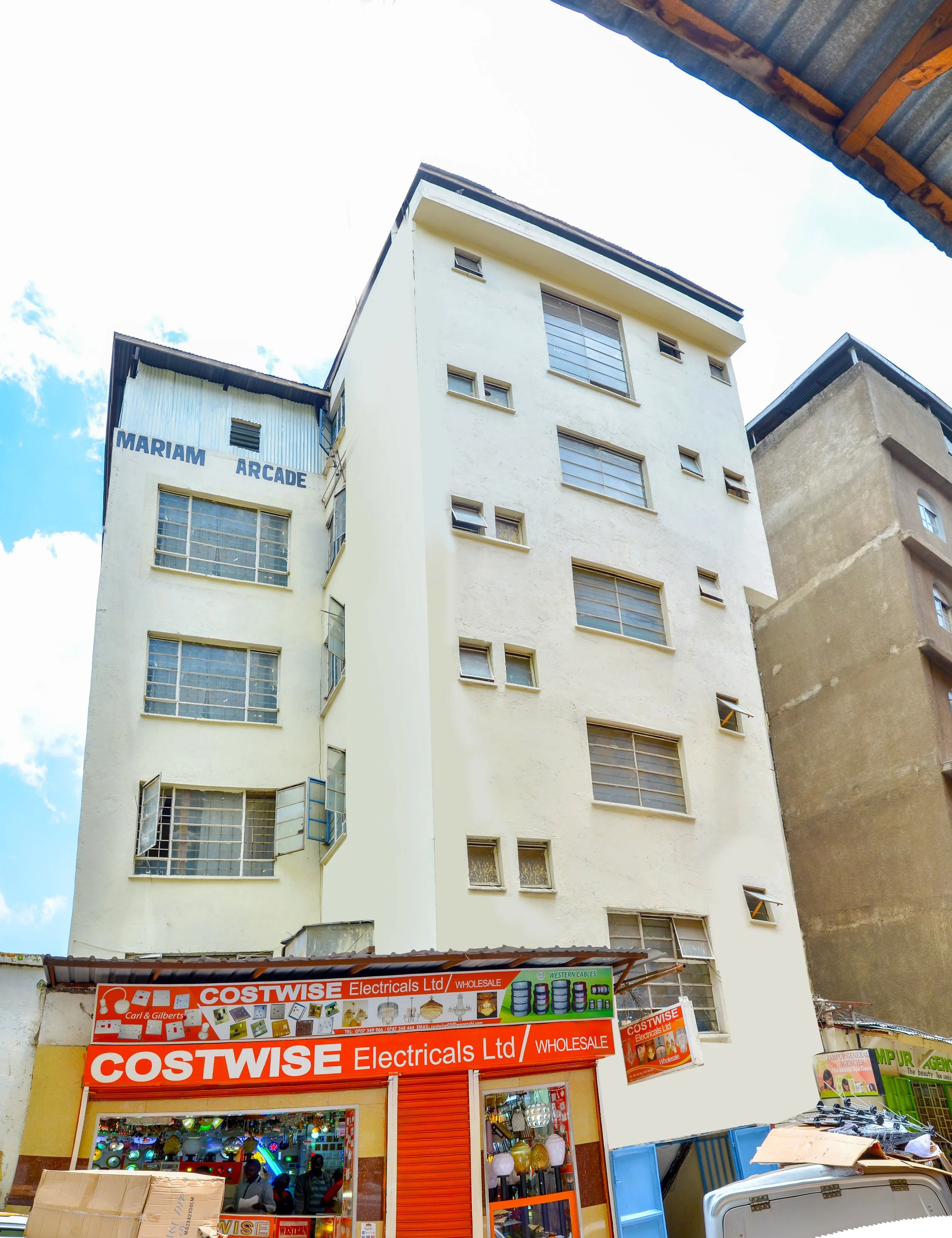 Tall, white building with multiple windows, signs reading "Mariam Arcade" and a shop called "Costwise Electricals Ltd" on the ground floor. The sky is partly cloudy; some objects and people are visible at street level.