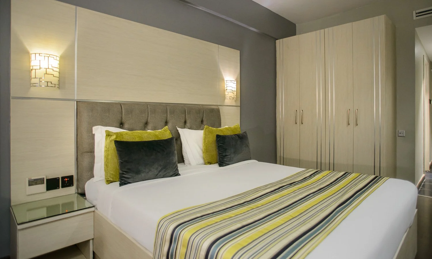 A neatly made hotel bed with a beige headboard, four pillows (two black and two green), and a striped bed runner in yellow, gray, and white. There is a nightstand with electrical outlets and a mirror, a beige wardrobe, and wall-mounted lamps on either side of the bed.