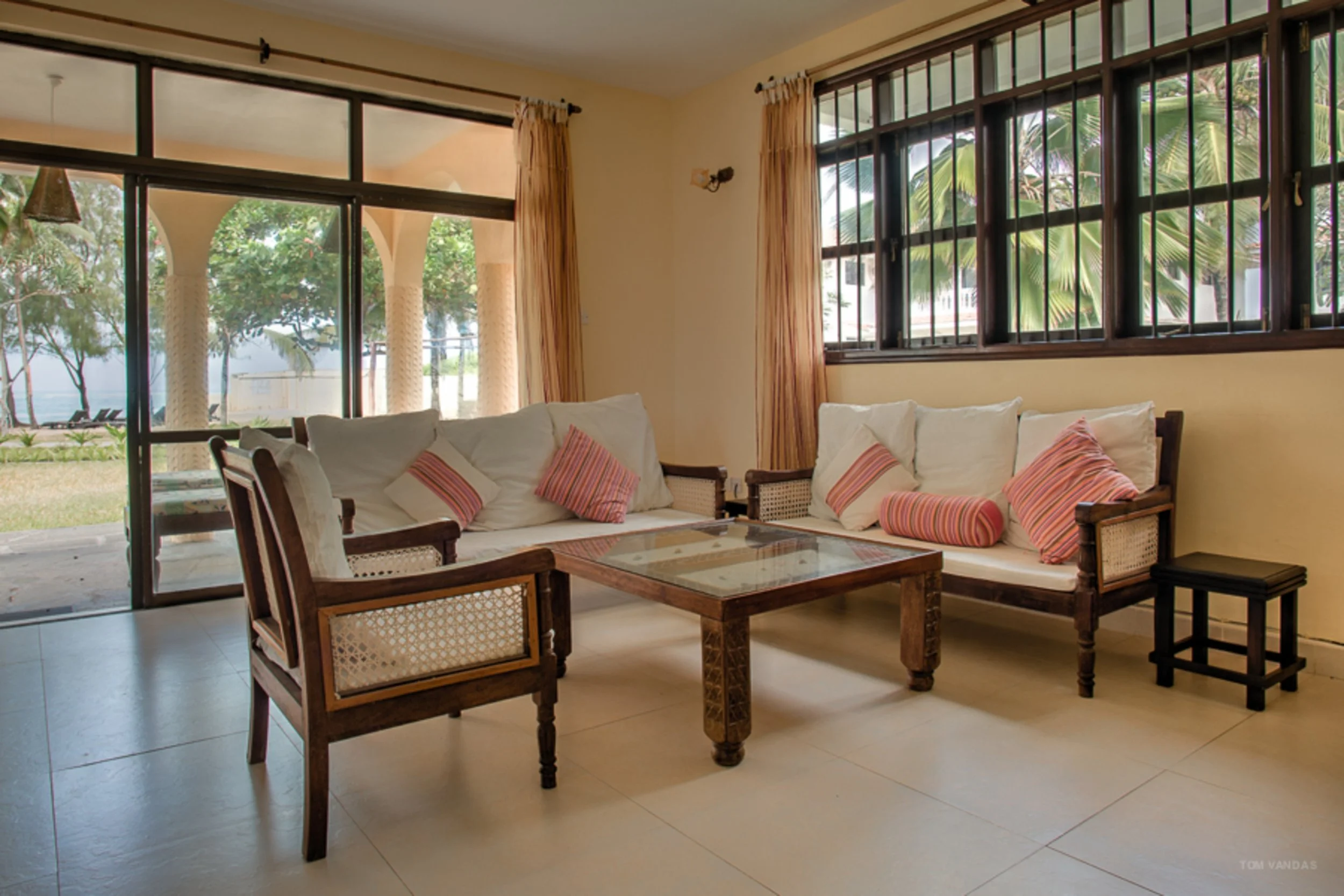 Living room with wooden furniture, white cushions, striped pillows, large windows showing palm trees outside, and a sliding glass door leading to an outdoor patio with greenery.