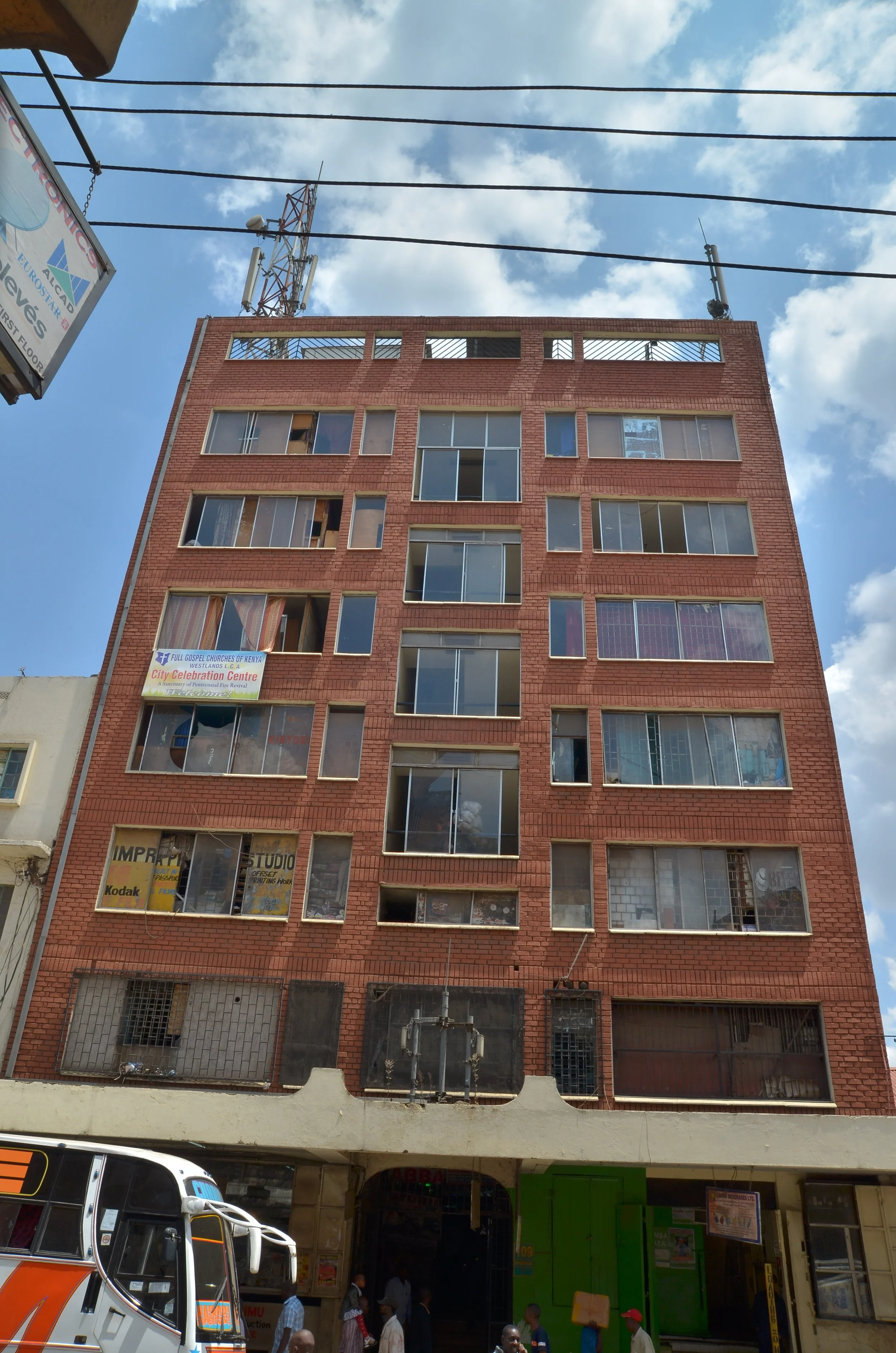 Multi-story brick building with several large windows, some with curtains or objects inside, and a sign promoting a city celebration center on one of the upper floors. The street level has a bus and people walking.