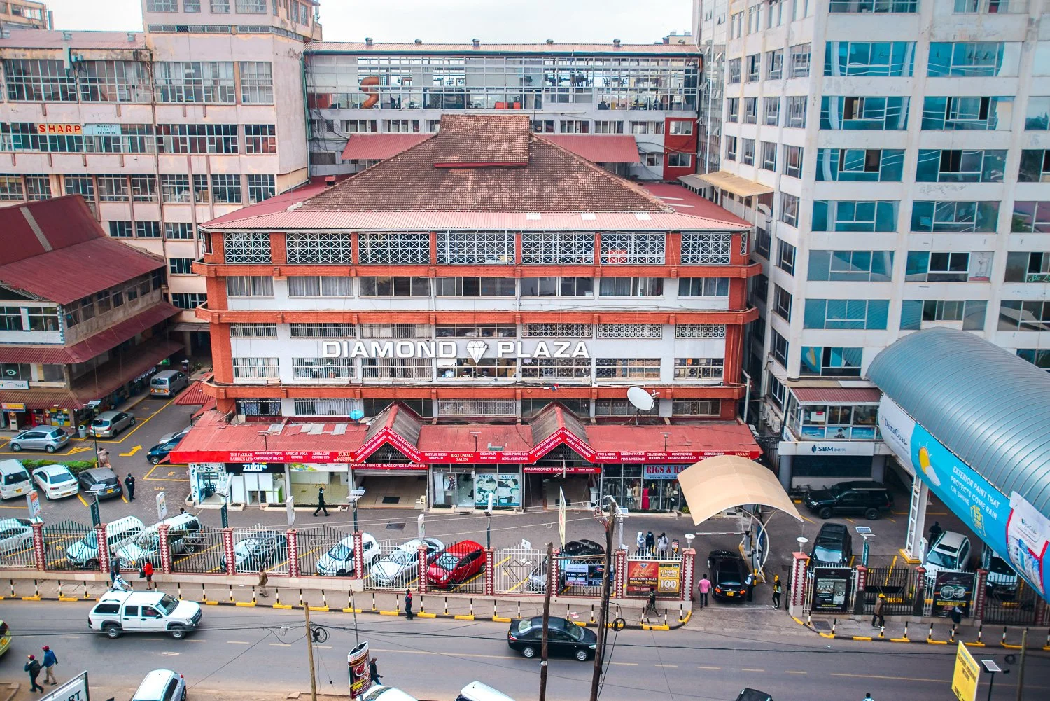 A multi-story commercial building called Diamond Plaza with shops on the ground floor and offices above, surrounded by parked cars and pedestrians, with tall office buildings on either side.