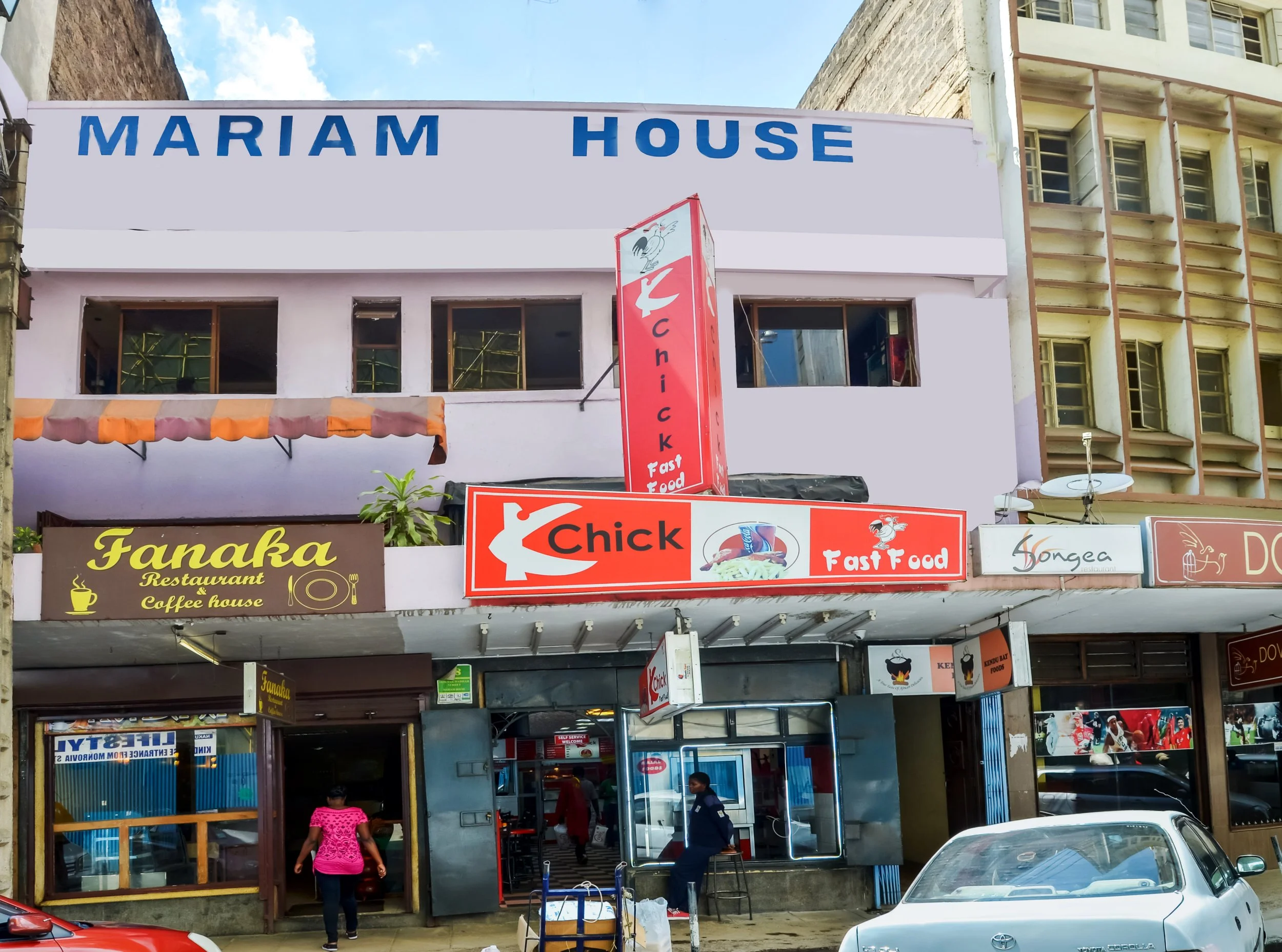 Street view of a building with signs for Mariam House, Fanaka restaurant, and Chick fast food, with people and cars in front.