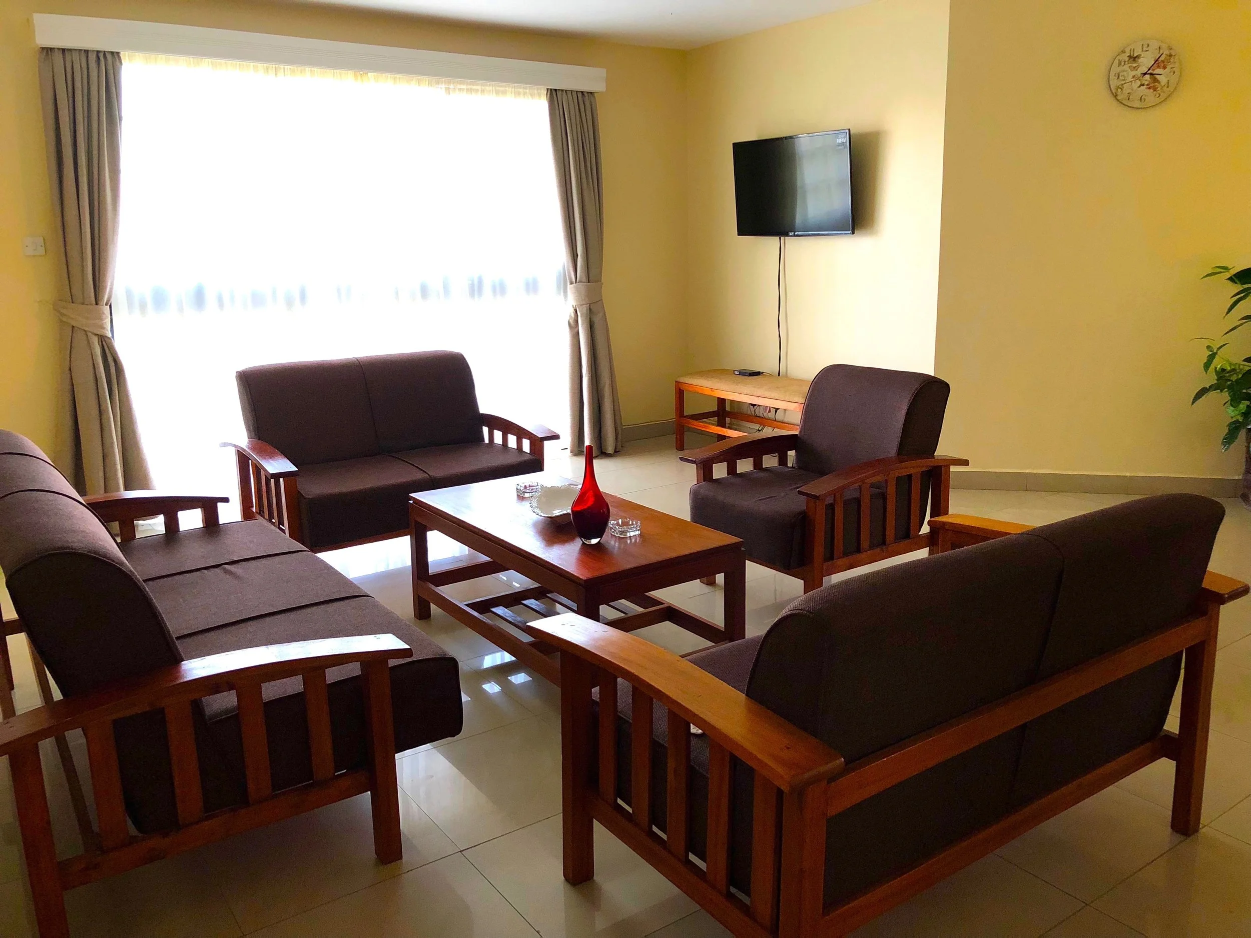 Living room with four brown cushioned sofas with wooden armrests arranged around a wooden coffee table, a large window with curtains, a wall-mounted TV, a small wooden side table, and a potted plant in the corner.