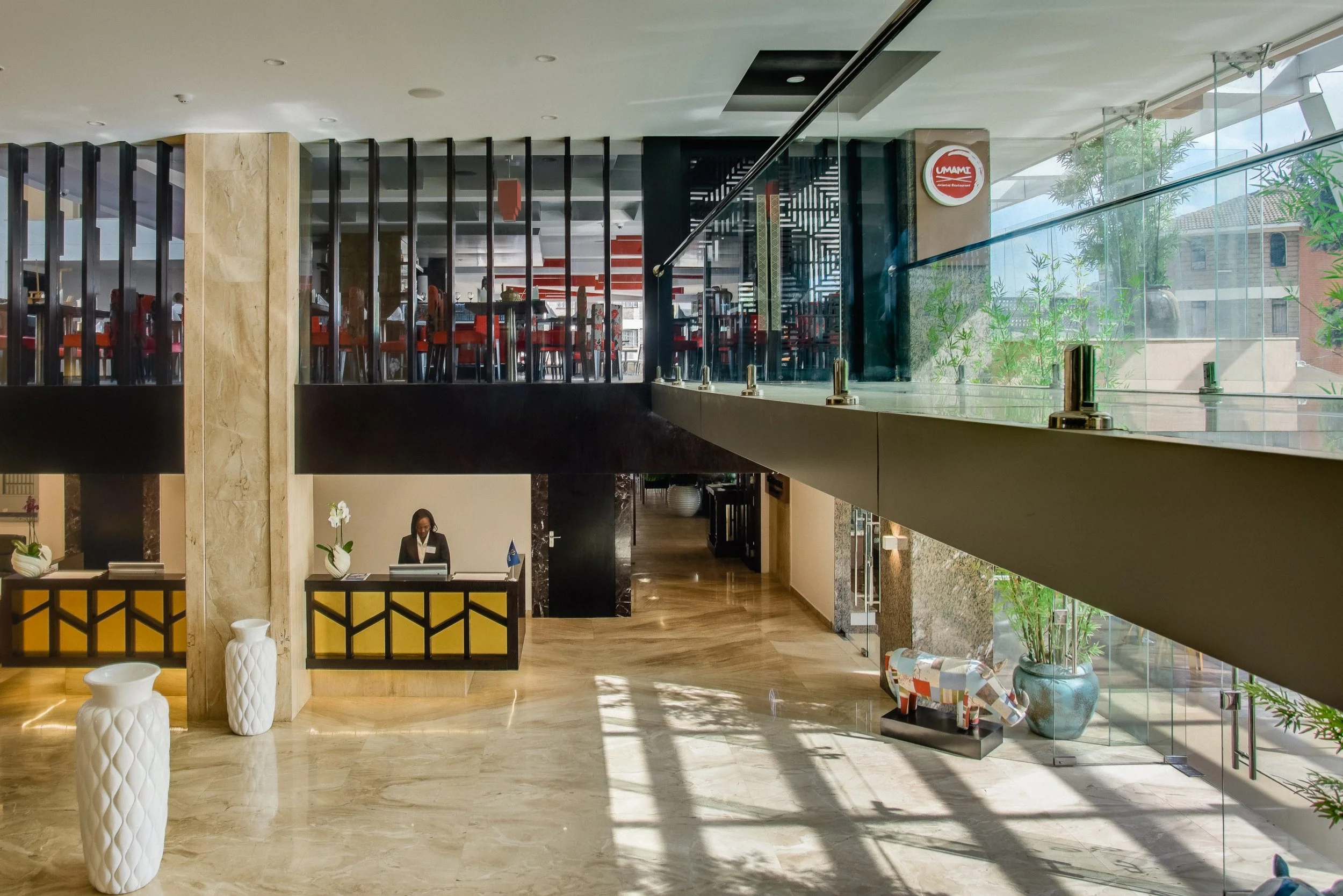 Hotel lobby with reception desk, a person working at the desk, glass railing, decorative vases, and potted plants, with sunlight streaming through large windows.