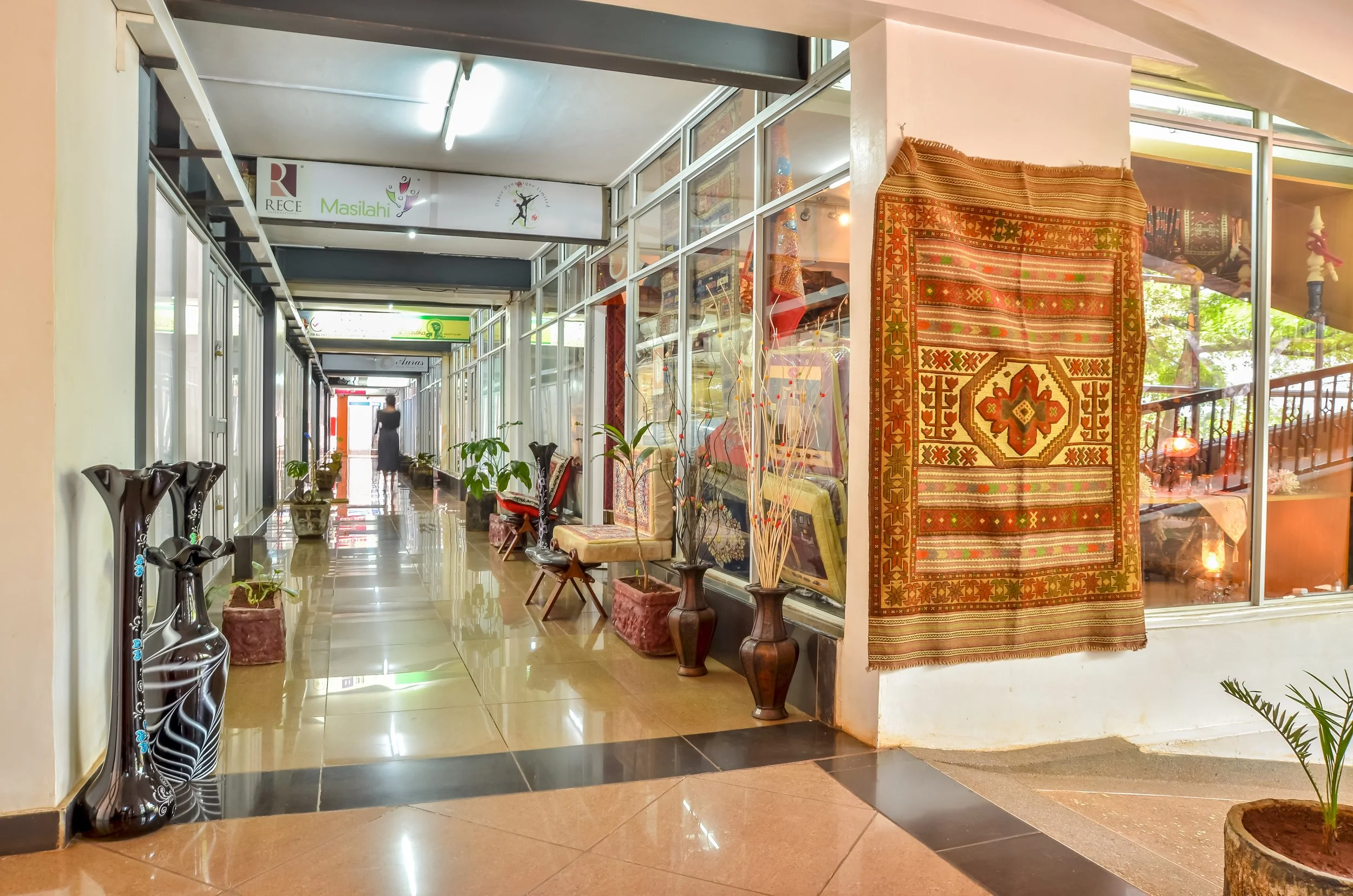 Indoor corridor with decorative vases, plants, and traditional textiles displayed in glass showcase.