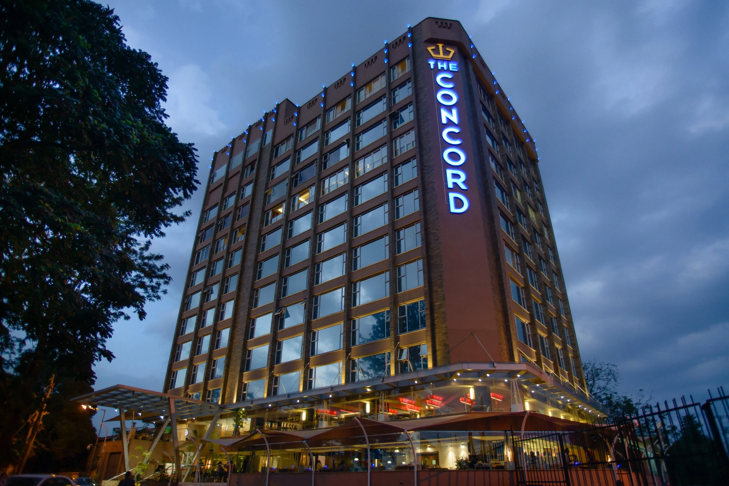 A tall hotel building named The Concord with illuminated blue neon sign at dusk, with trees and cloudy sky in the background.