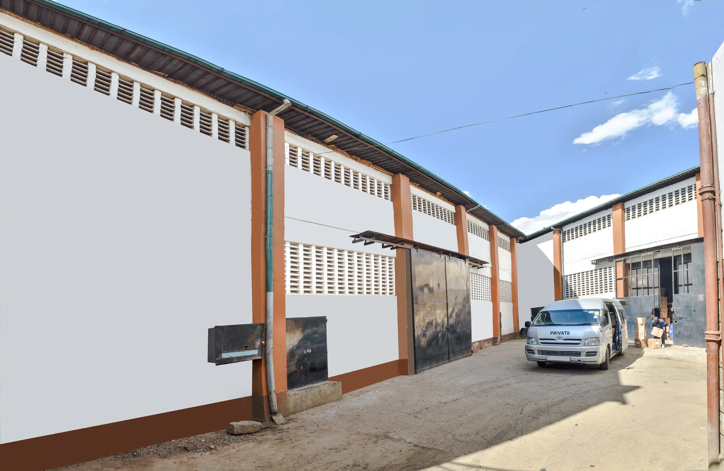 An industrial building with a white wall, orange accents, and metal gates. A white van marked 'Private' is parked outside, and a person is seen unloading boxes nearby. The ground is unpaved, and the sky is clear with a few clouds.