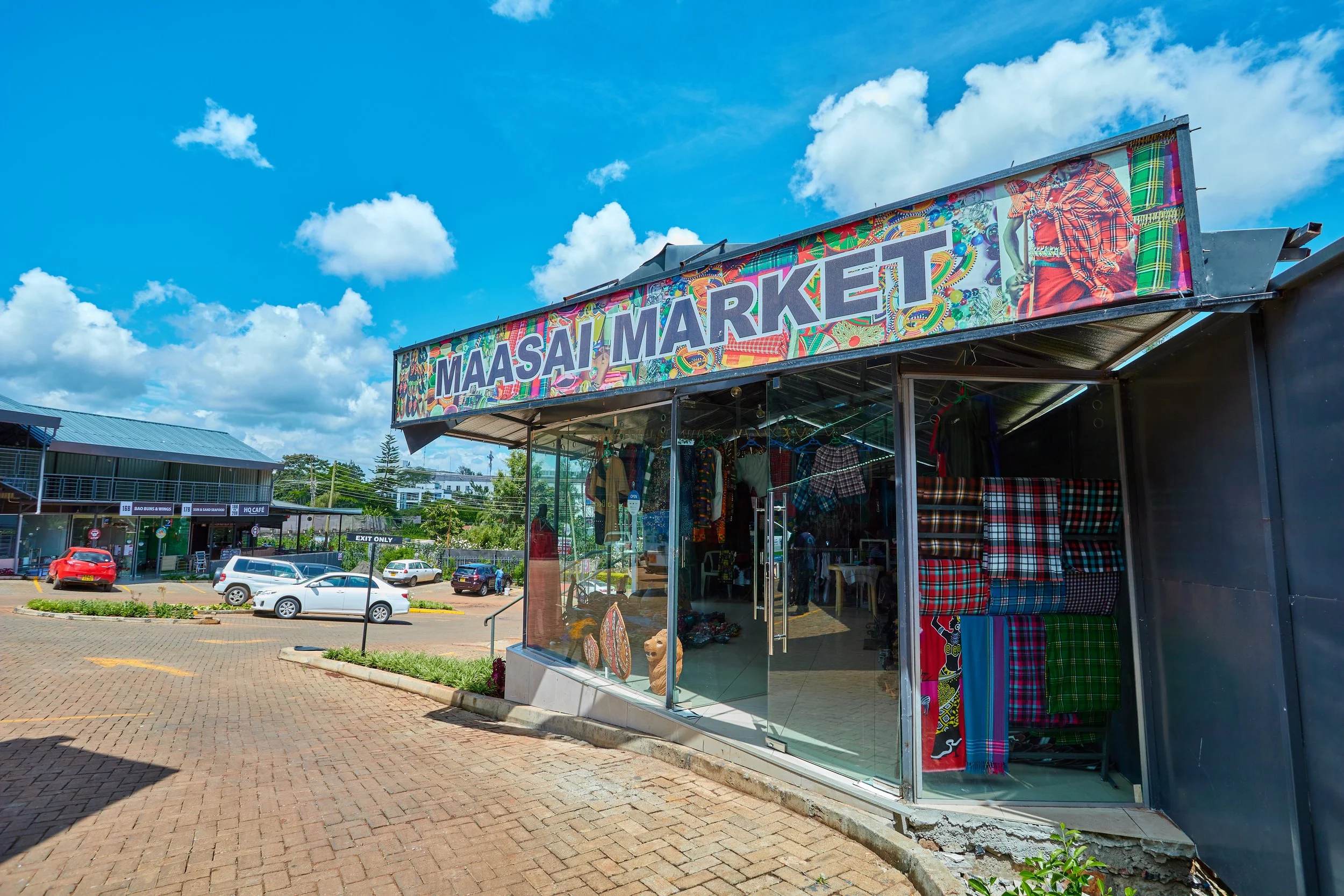 Exterior view of Maasi Market shop with colorful clothing and fabric displays, parking lot with several cars, blue sky with scattered clouds.