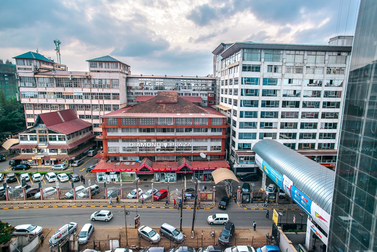View of a busy city street with multiple buildings, parked cars, pedestrians, and a large sign for Diamond Plaza in the center. Overcast sky overhead.