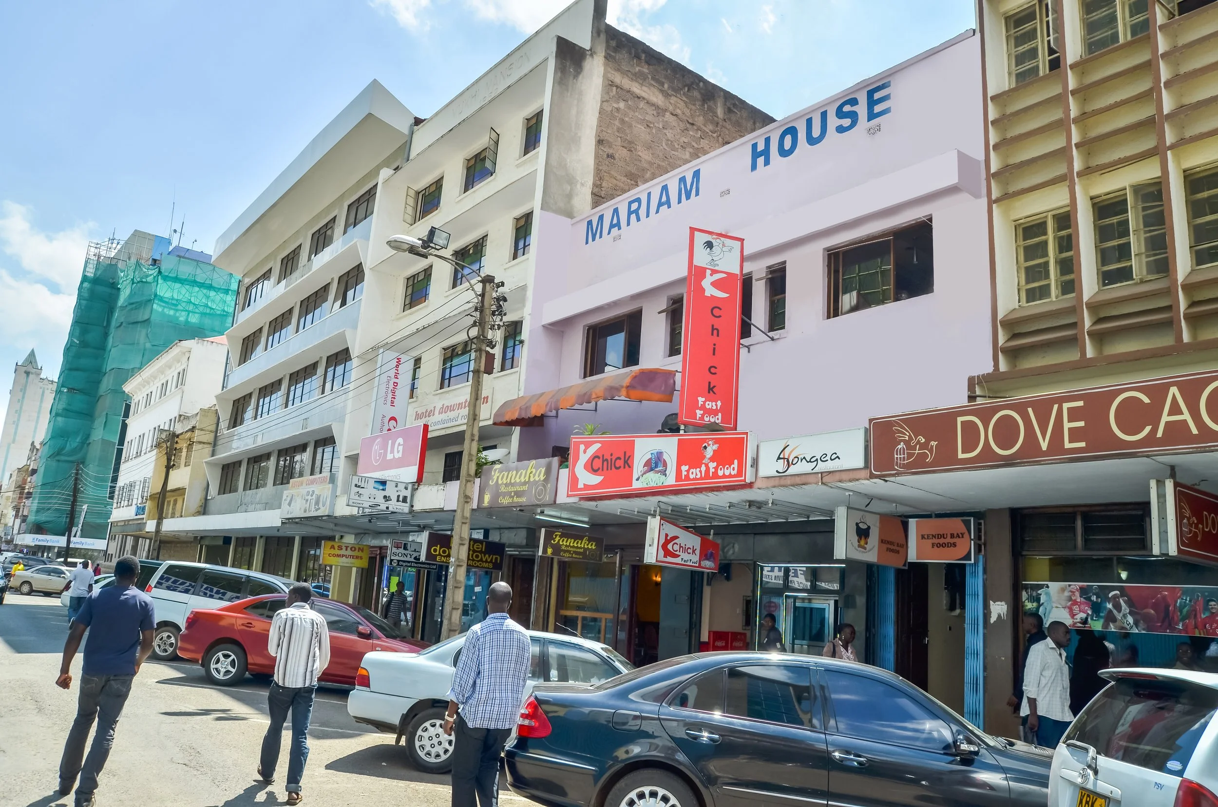 Street view of commercial buildings with signs for restaurants, shops, and offices; cars parked along the street; people walking on the sidewalk.