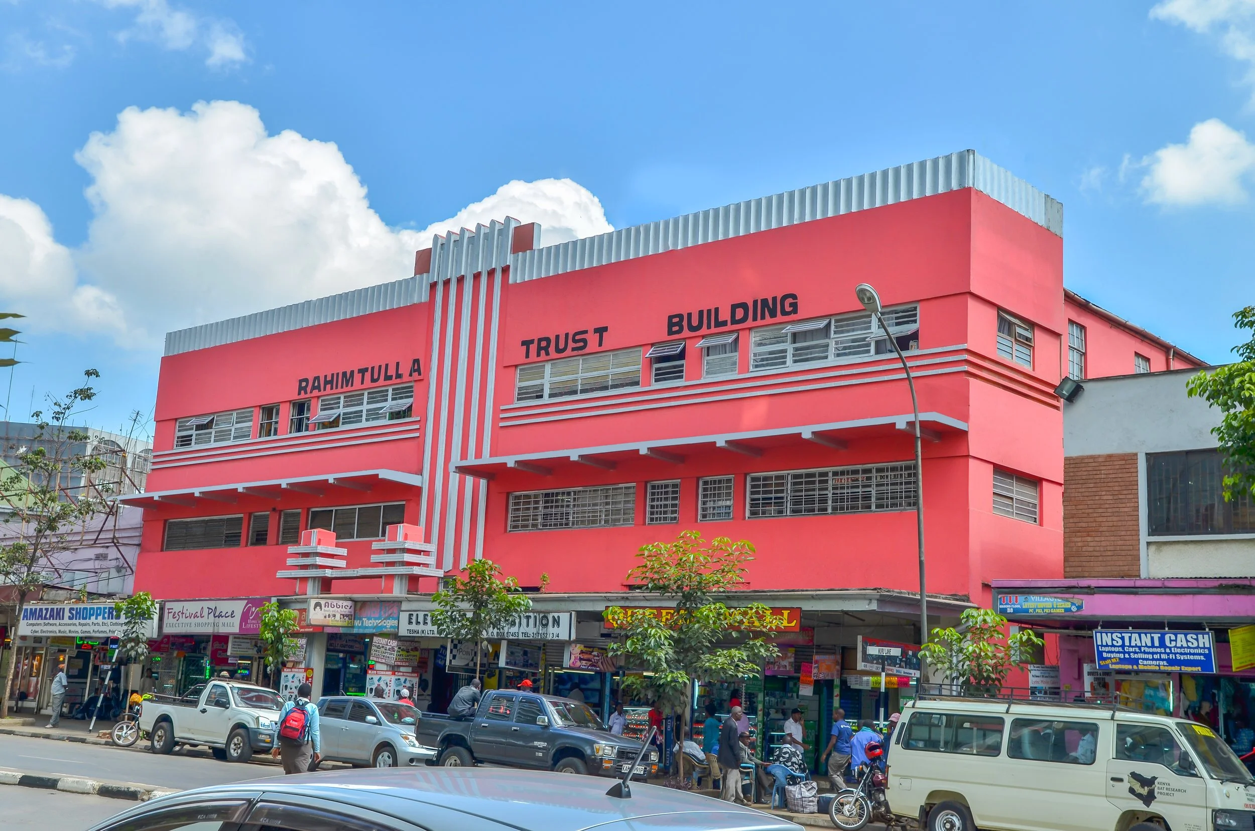 A pink multi-story building with a sign that reads 'Rahimtulla Trust Building'. The ground floor has shops and people walking on the street, and there are cars and vans parked in front.