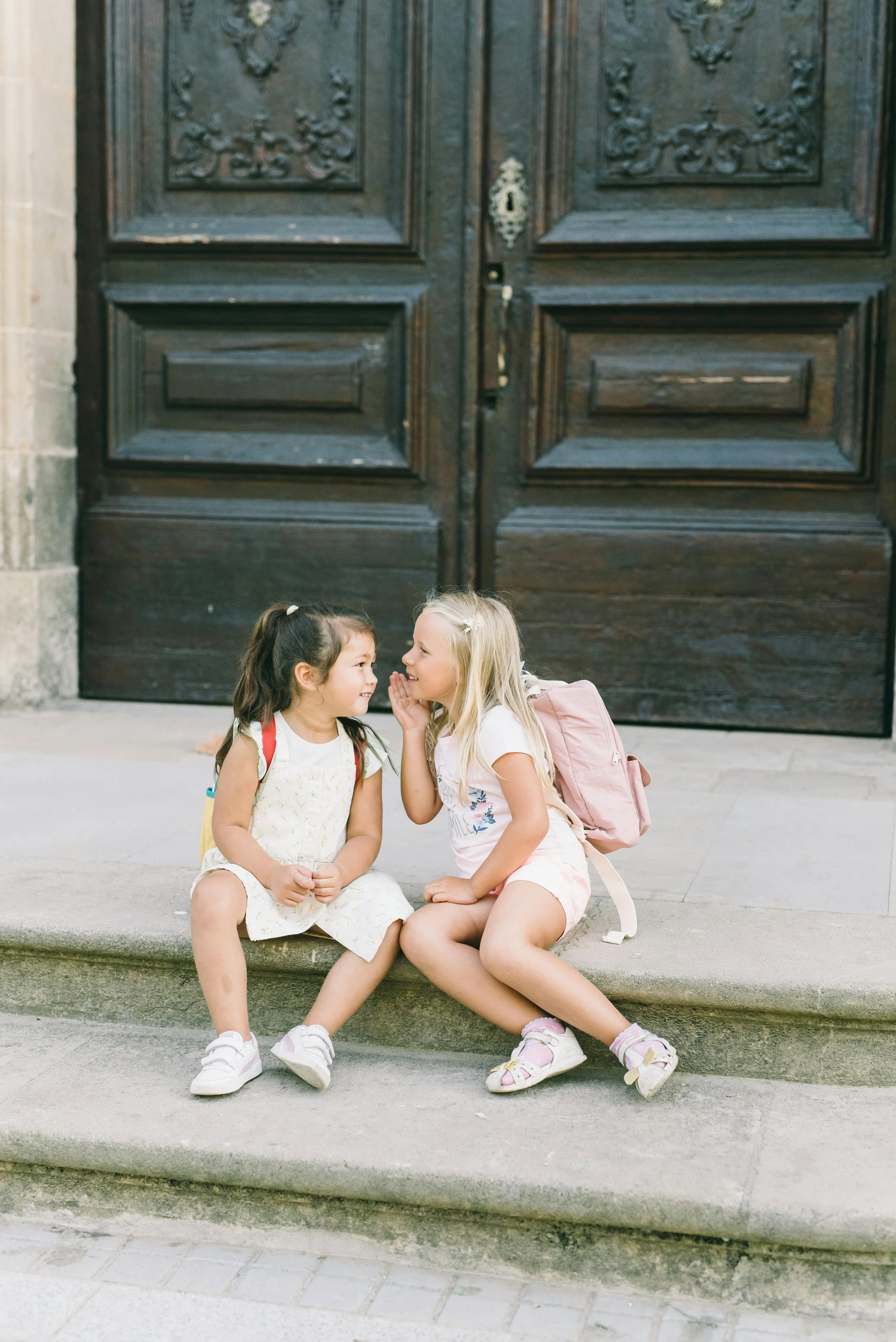 Two young girls sitting on stone steps in front of a large wooden door, engaging in a secret conversation, both wearing backpacks and casual clothing.