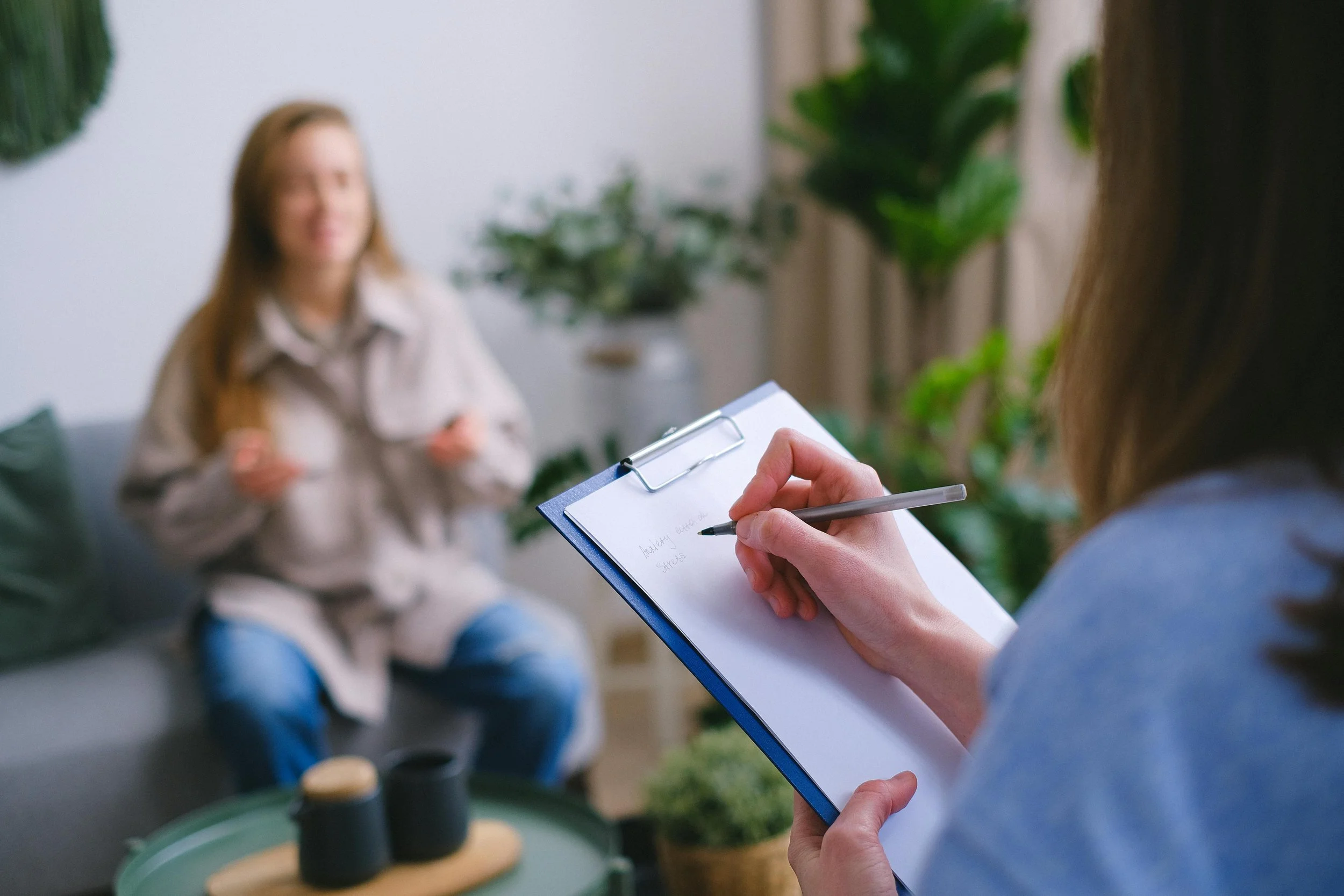 A woman taking notes on a clipboard during a therapy session with a smiling woman sitting on a couch in a living room with plants.