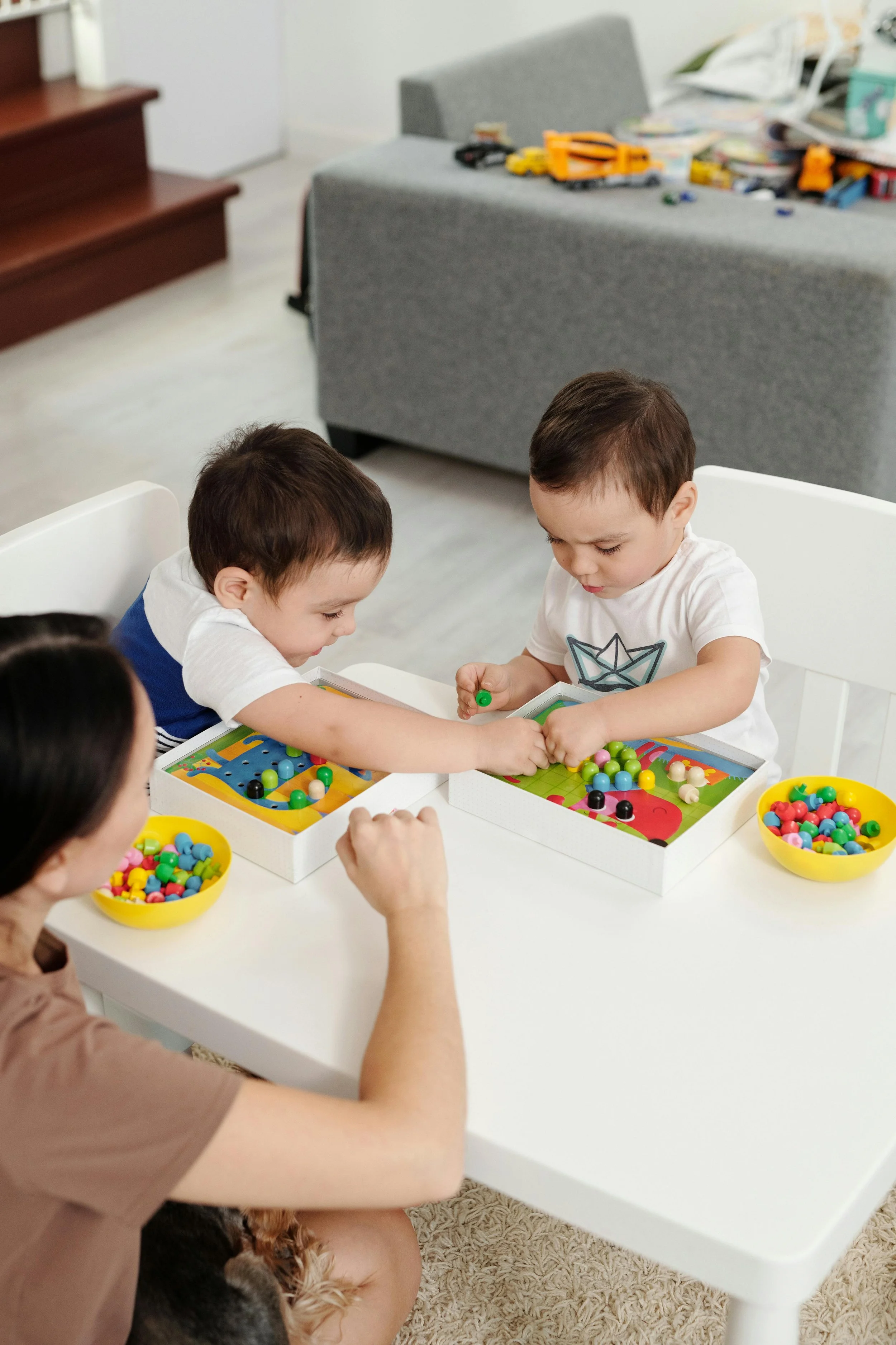 Two young boys and a woman playing with a board game at a white table. The boys are focused, and there are bowls of colorful game pieces on the table. The background shows a gray couch and a wooden staircase.
