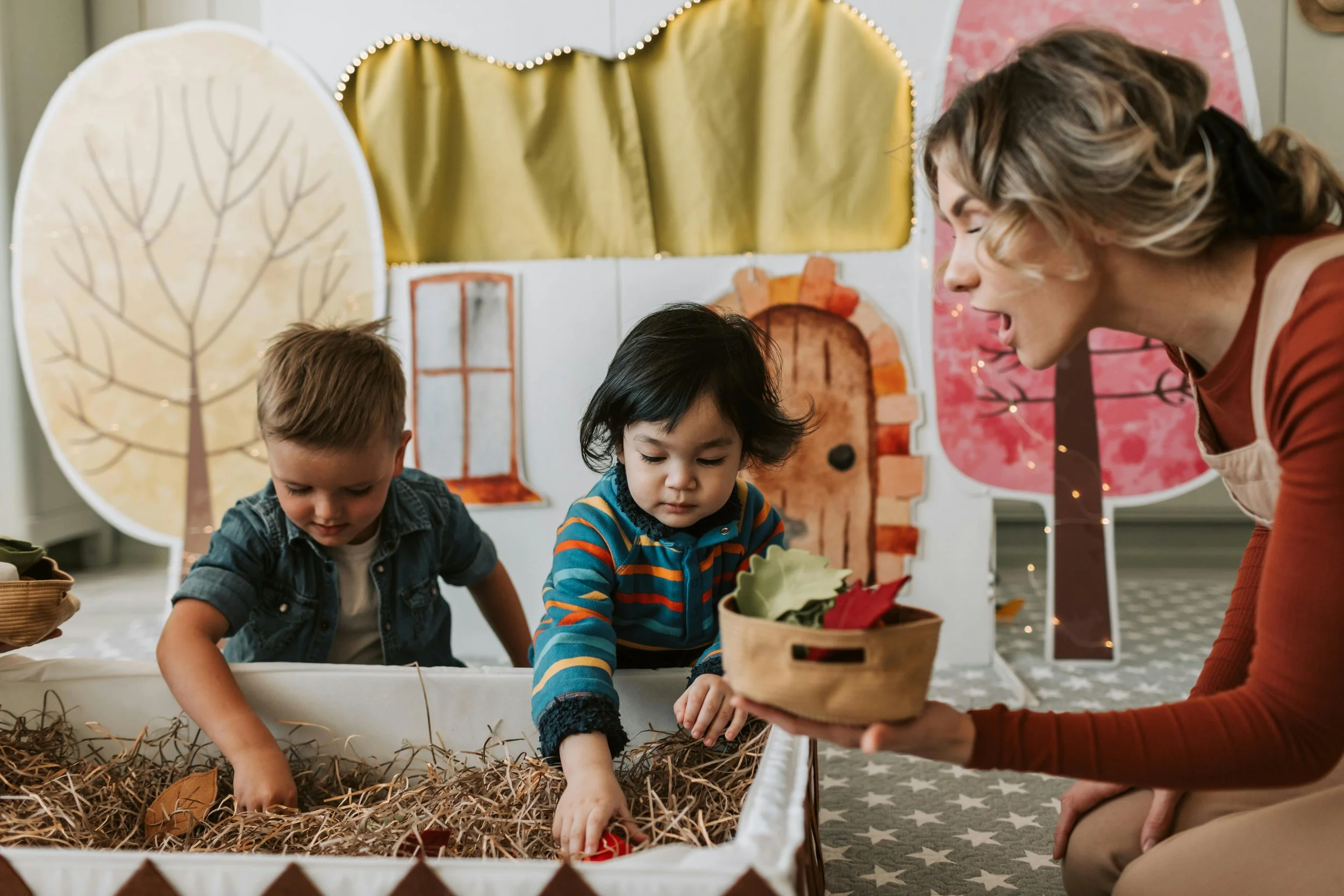 A woman is kneeling next to two children, engaging in a pretend play activity with autumn leaves in a box, in a setting decorated with colorful paper trees and a house backdrop.