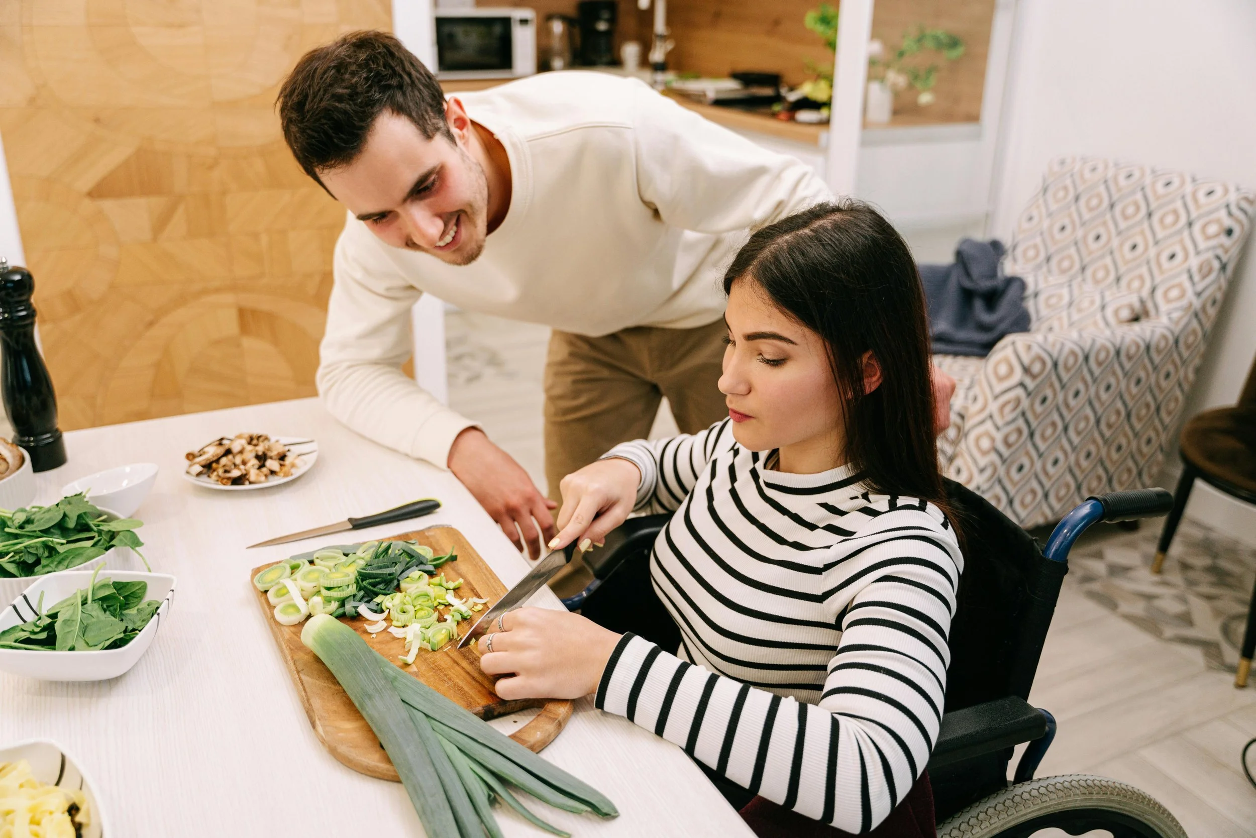 A woman in a wheelchair chopping green onions at a table, with a man smiling and leaning over her, in a cozy kitchen setting.