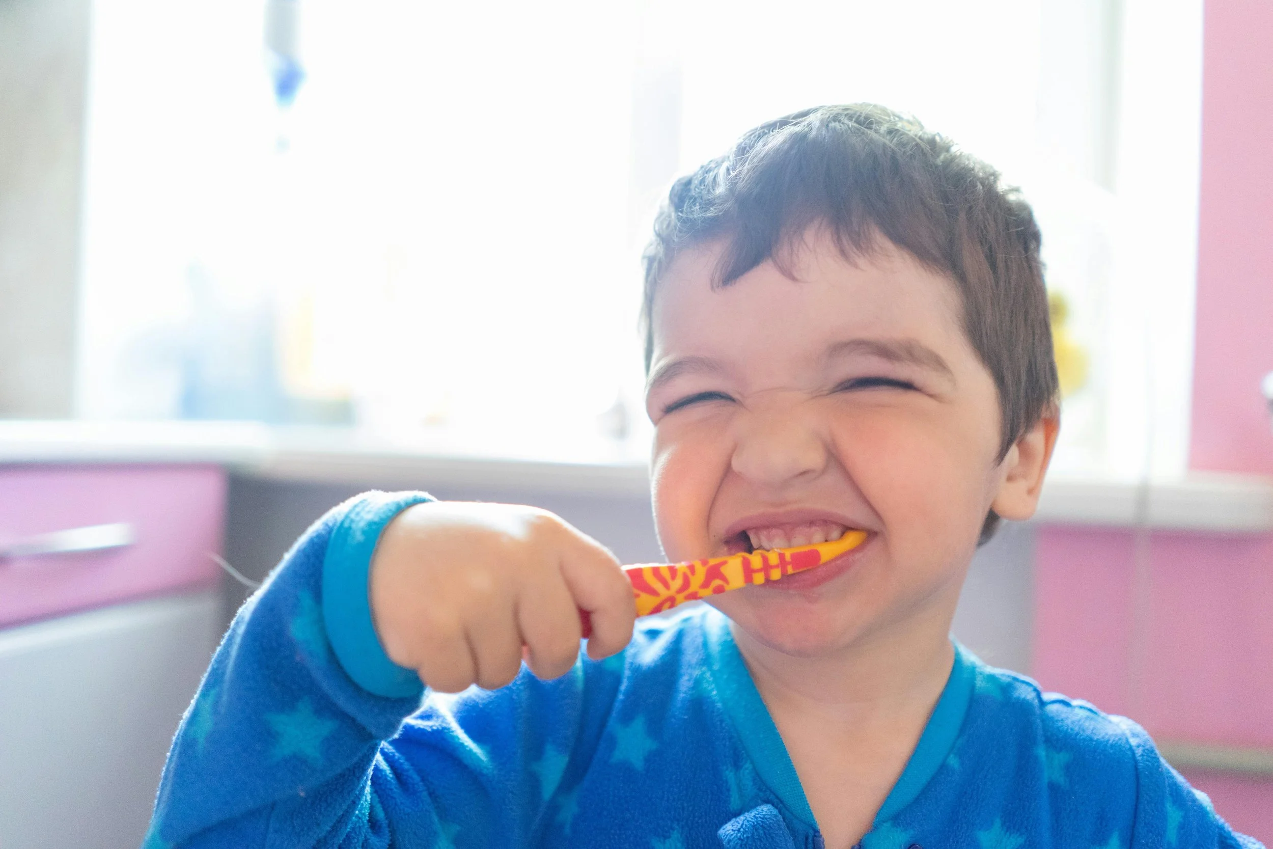 A young boy with dark hair smiling as he bites on a colorful toothbrush, wearing a blue shirt, in a brightly lit room with pink and white cabinets and a window in the background.