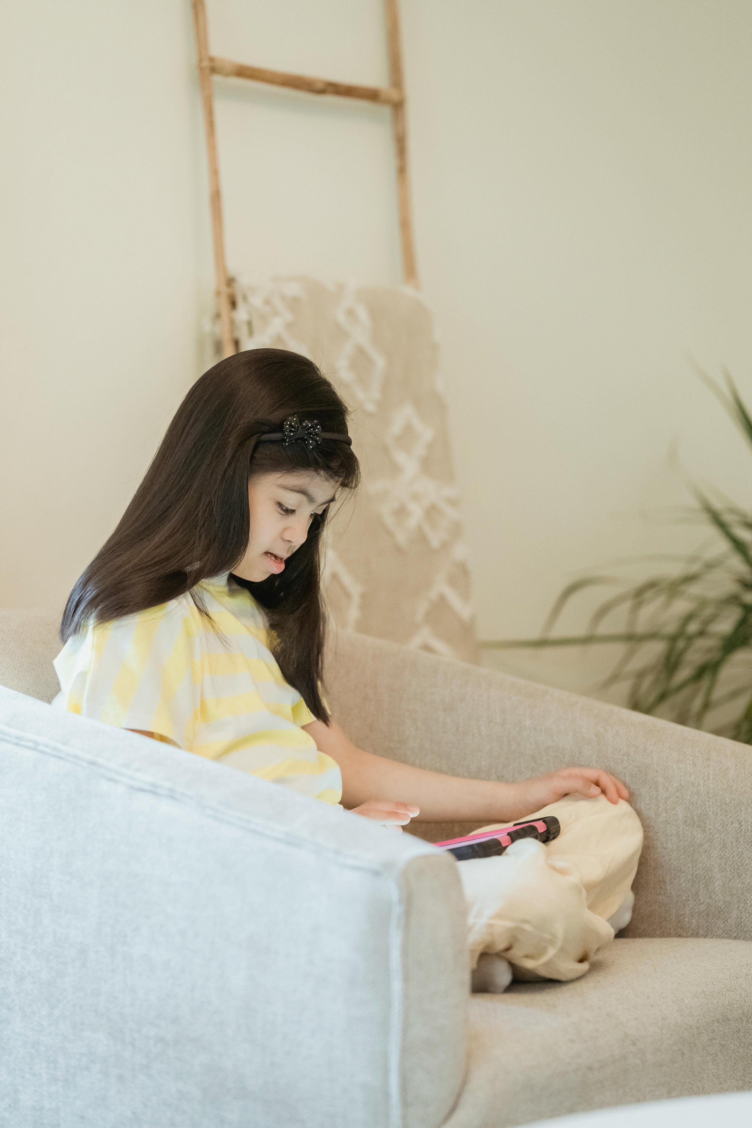 A young girl with long dark hair, wearing a yellow and white striped shirt, is sitting on a beige sofa. She is looking down at and touching a tablet or electronic device in her lap, with a concentrated expression. In the background, there is a decorative ladder against the wall with white lace fabric hanging on it and some indoor plants.