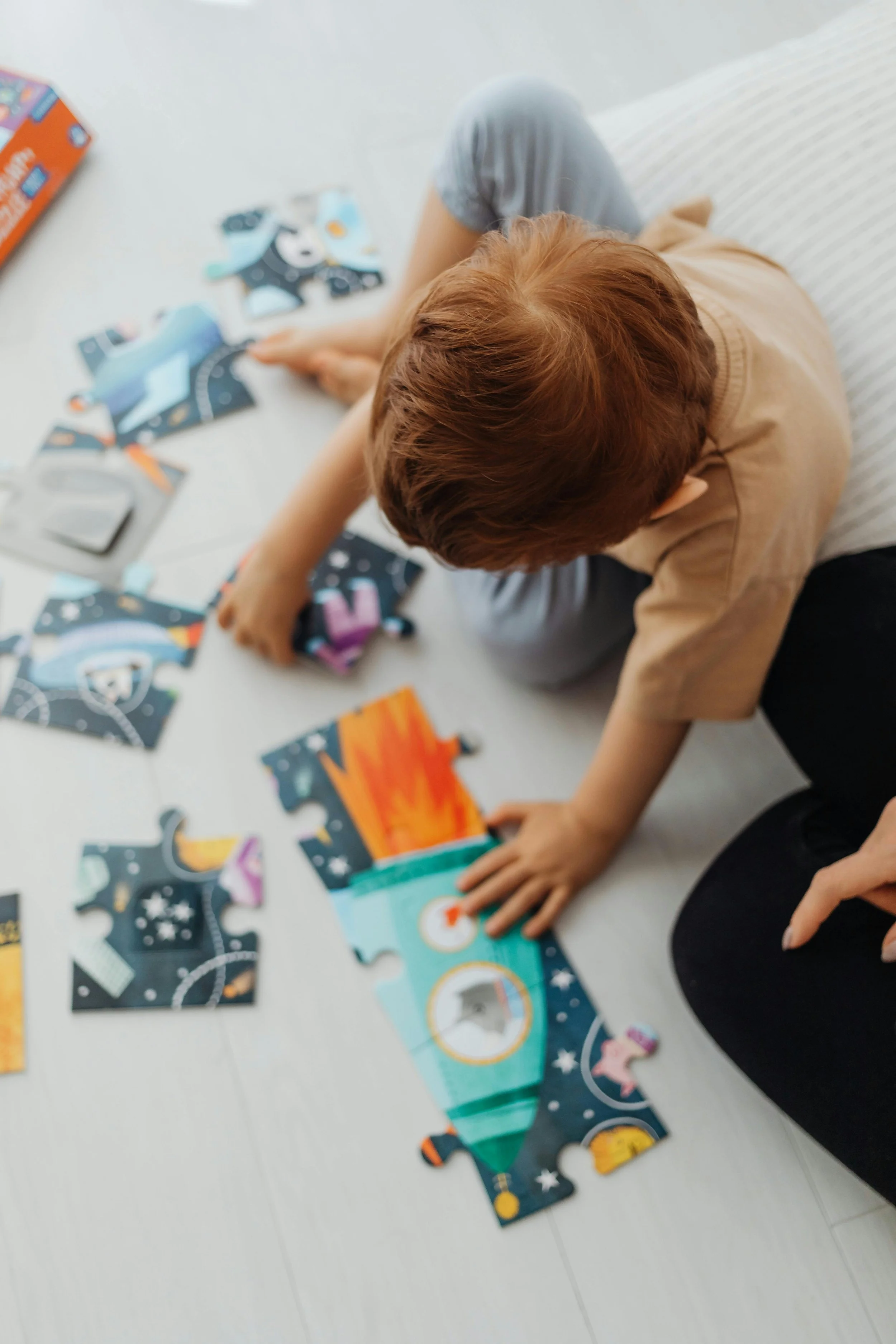 A child with reddish hair sitting on a light-colored floor, assembling a colorful space-themed jigsaw puzzle with an adult's hand pointing at it. The puzzle features a rocket and outer space design.