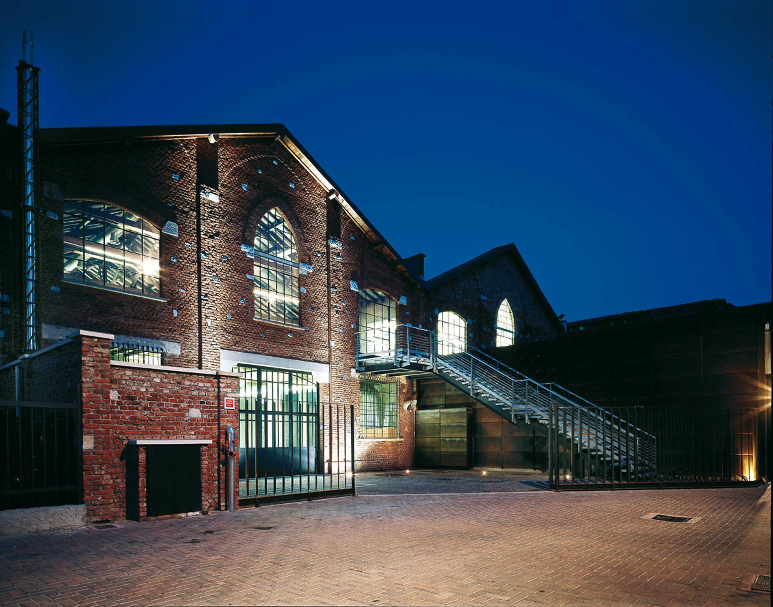 Edificio in mattoni rossi con grandi finestre, illuminato di sera, con una scala esterna in metallo e un cancello di ferro.
