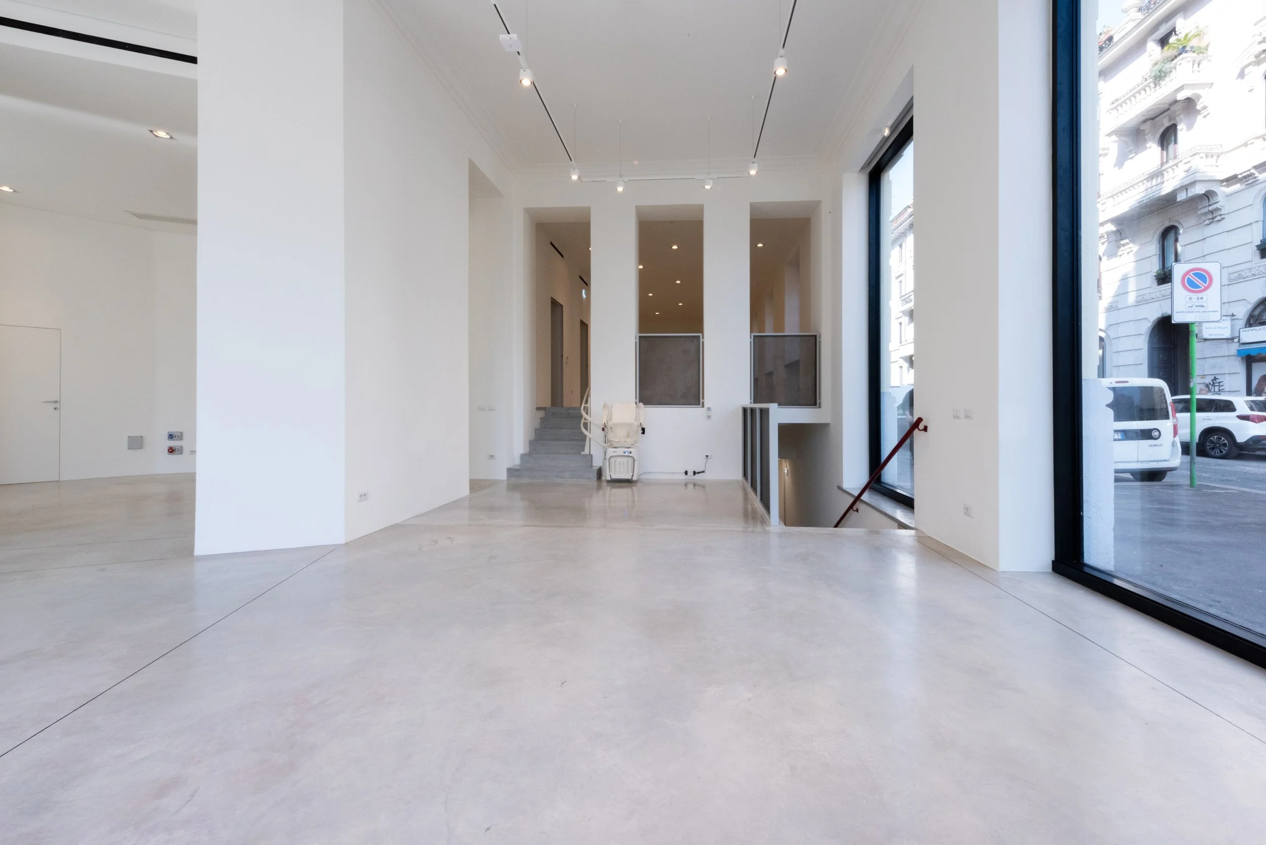 Interno di un edificio con pareti bianche, grandi finestre e pavimento in cemento, attualmente non arredato.