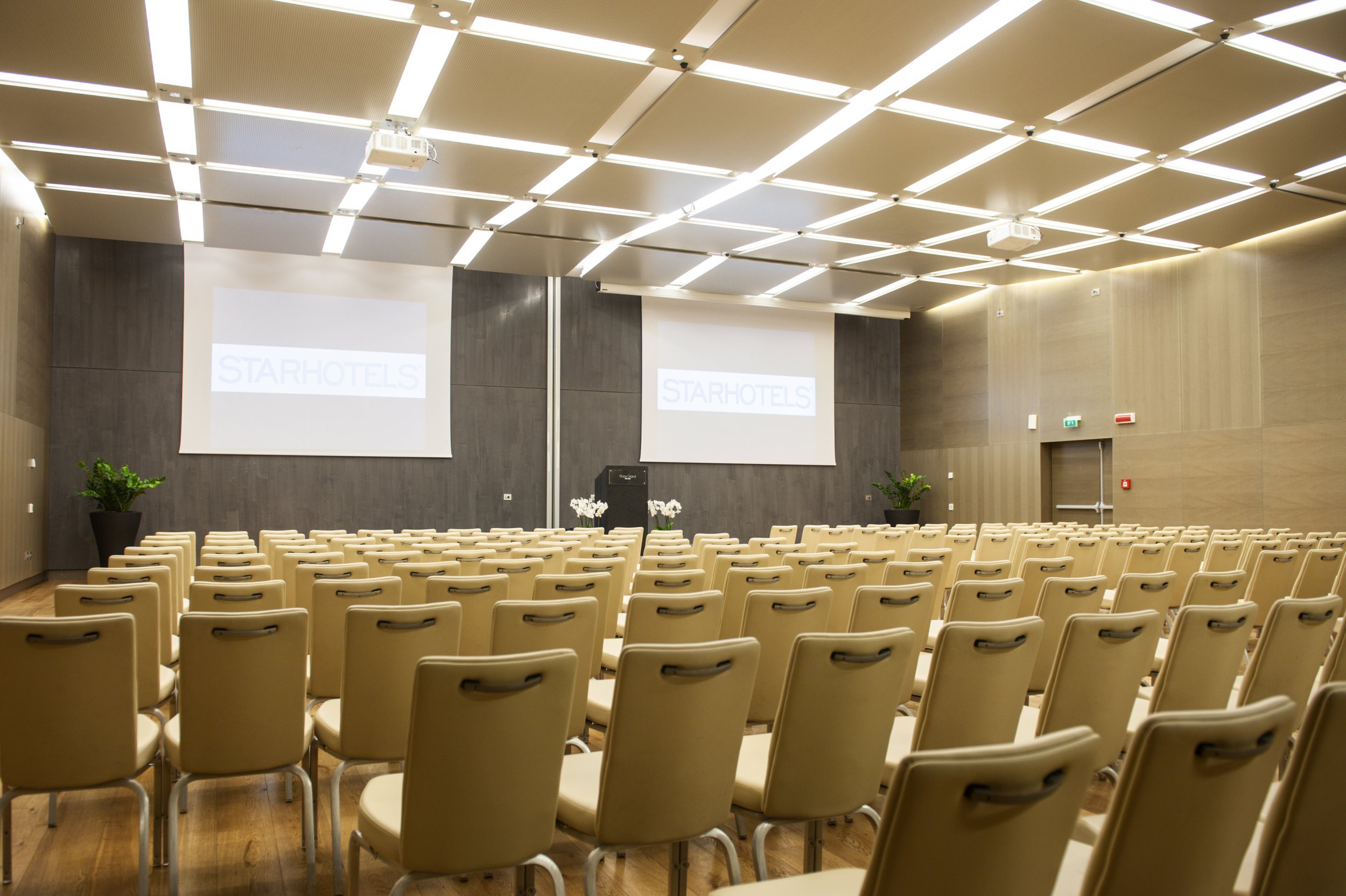 Sala conferenze con sedie beige, due schermi con il logo 'Starhotels', e piante verdi, arredamento moderno