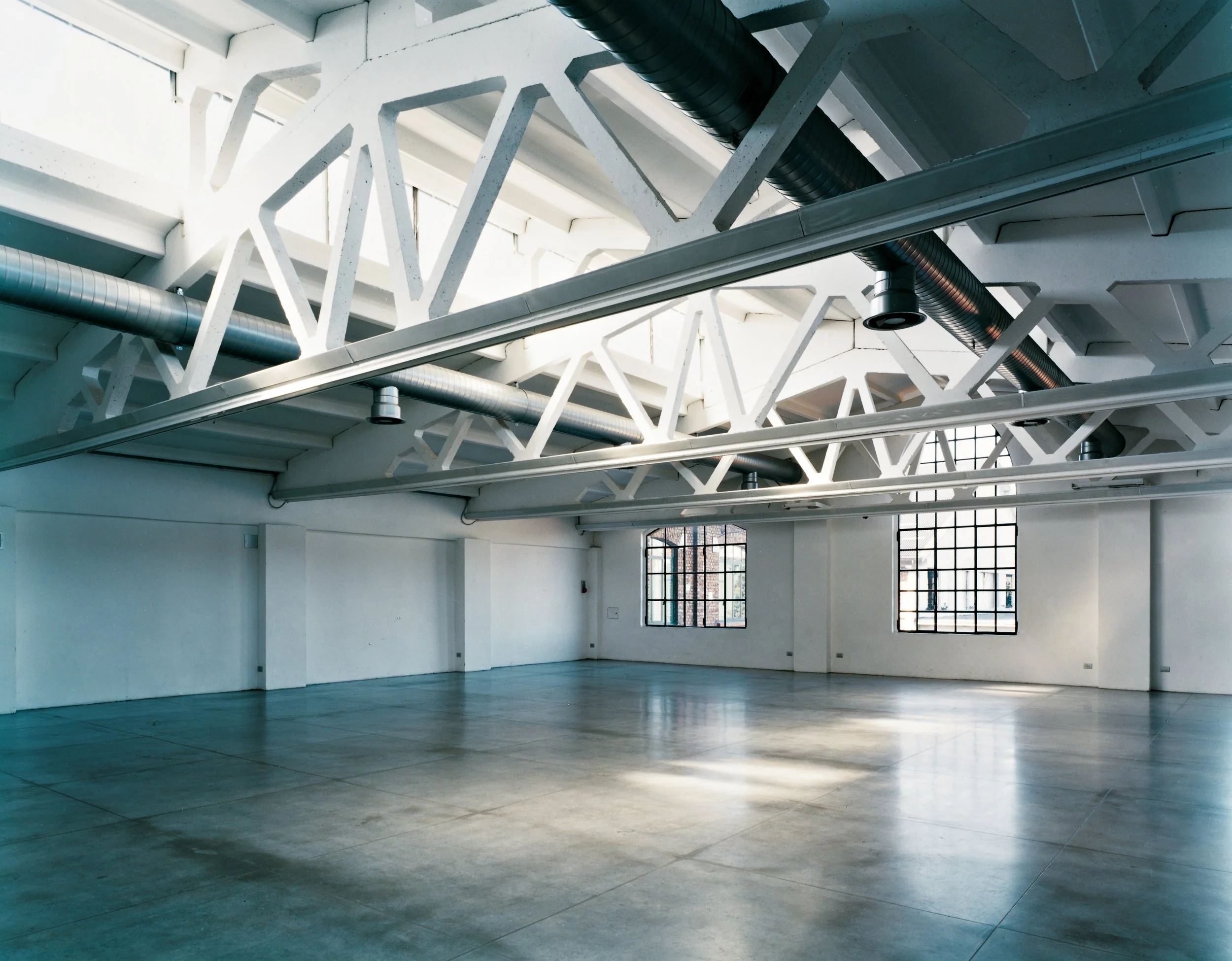 Interno di un grande spazio vuoto con pareti bianche, grandi finestre e travi e condotti a vista sul soffitto.