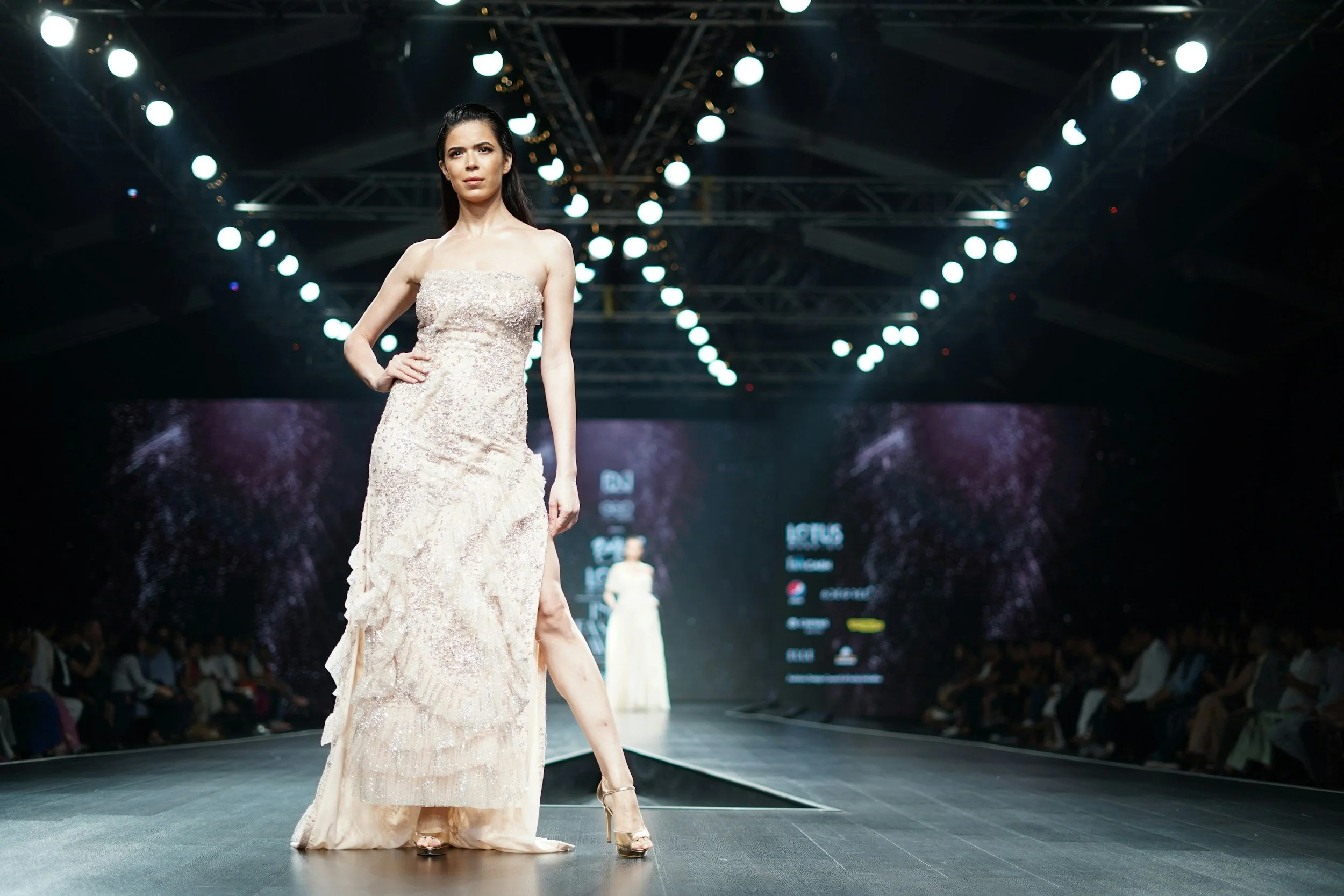 Una modella indossa un abito da sera lungo e sobrio, cammina lungo una passerella in un evento di moda, sotto luci intense e un grande schermo digitale sullo sfondo.