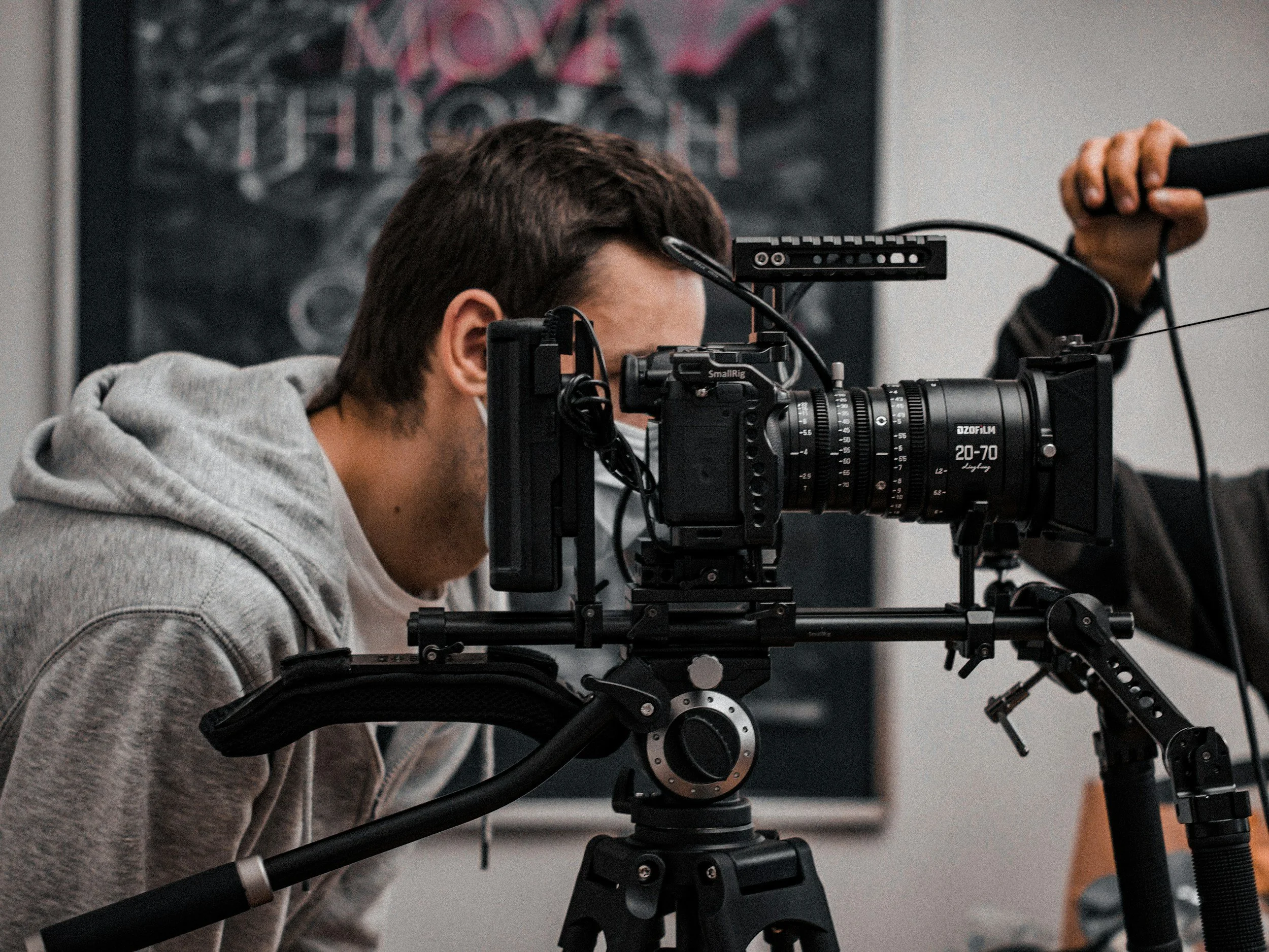A young man is operating a professional film camera on a tripod, wearing a gray hoodie and face mask, in an indoor setting.