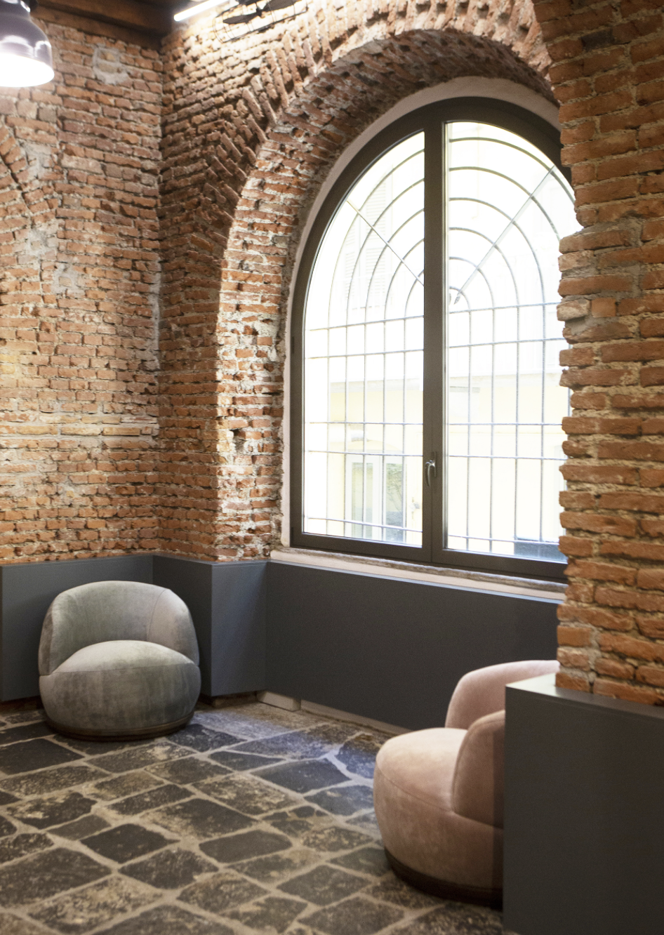 Interno di una stanza con pareti di mattoni rossi e una grande finestra ad arco con inferriate, due sedie imbottite di colore beige e grigio, pavimento in pietra