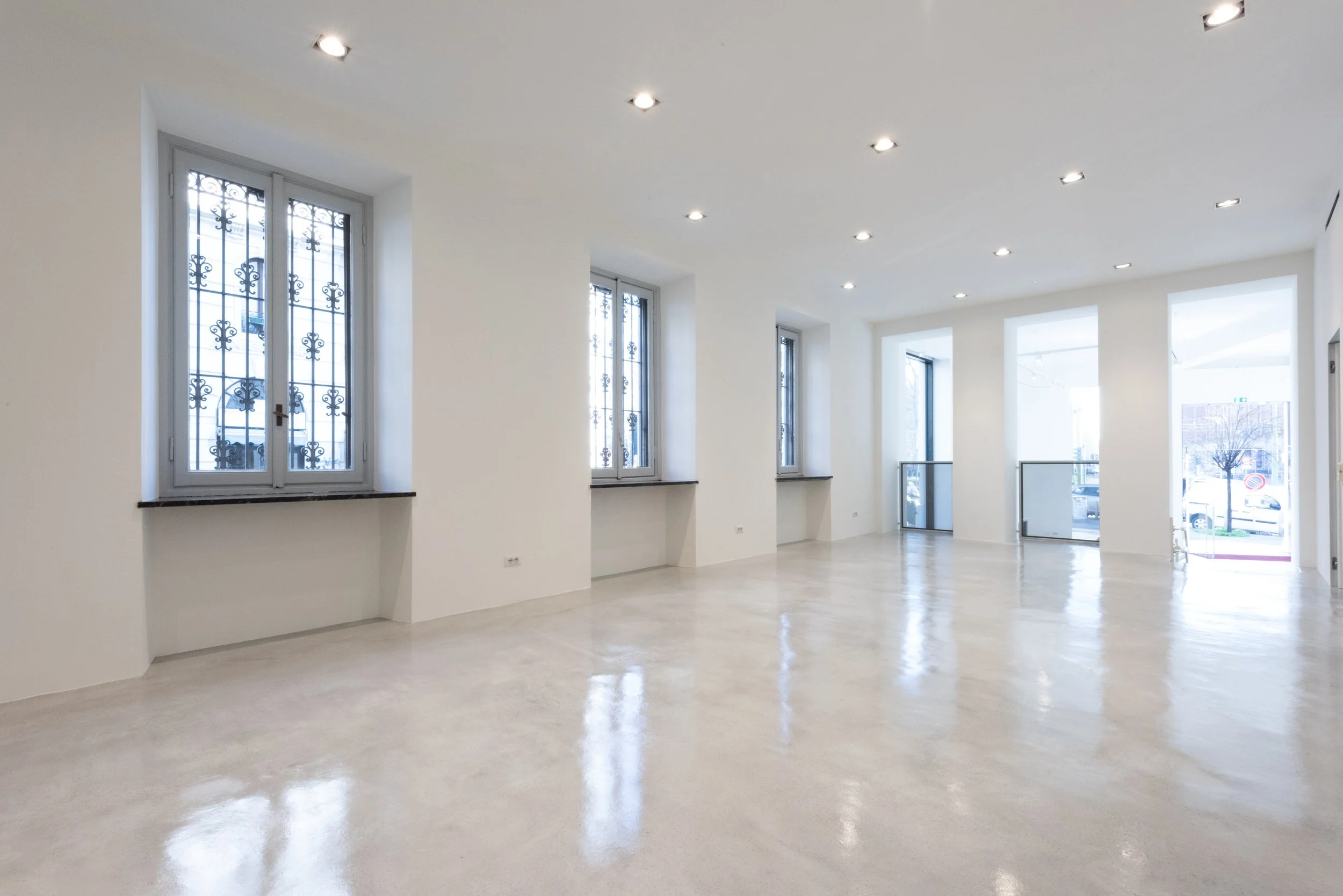 Interno di un locale vuoto con pareti bianche, grandi finestre con grate e illuminazione a soffitto.