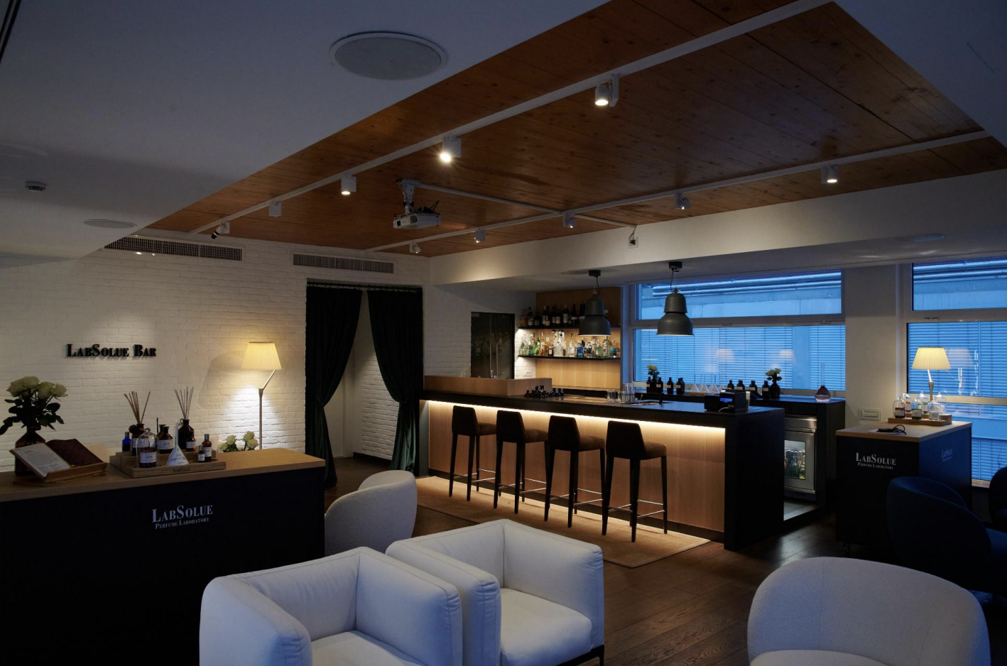 interni di un bar elegante con sedie nere e un bancone in legno illuminato, divani bianchi e decorazioni con piante e lampade