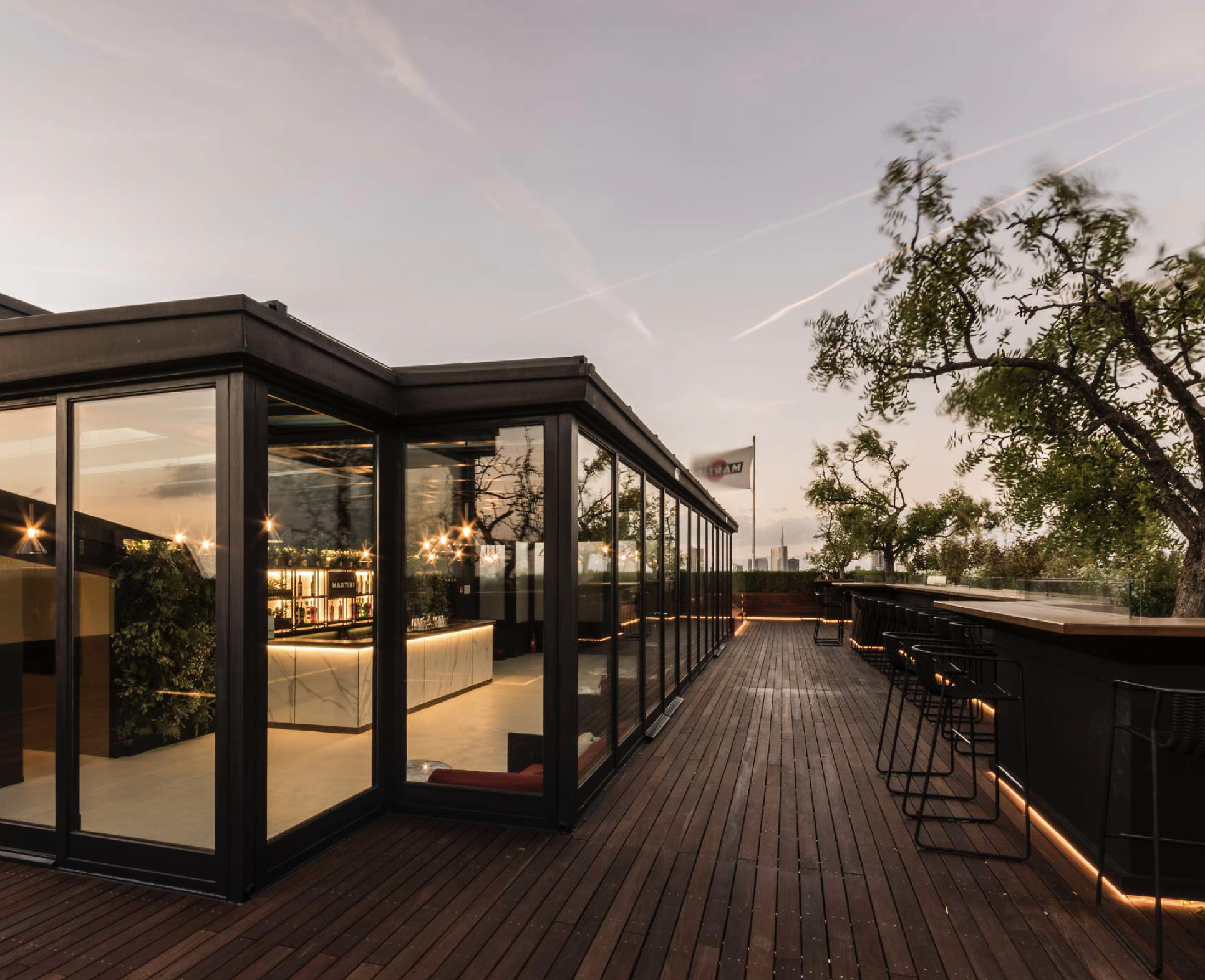 Terrazza moderna con bar all'aperto e grandi finestre che riflettono le luci interne, con tavoli e sedie nere e un cielo al tramonto.
