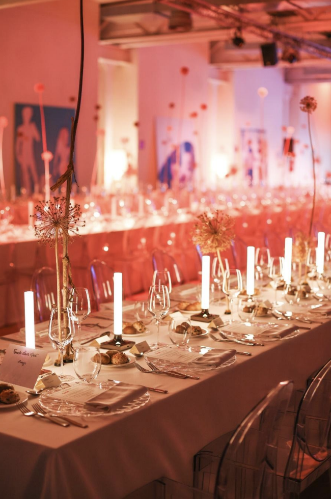 Tavola appare elegantemente preparata con bicchieri, piatti, posate, candelabri con candele accese e fiori decorativi, in un ambiente decorato per un evento o festa, con luci calde e decorazioni sullo sfondo.